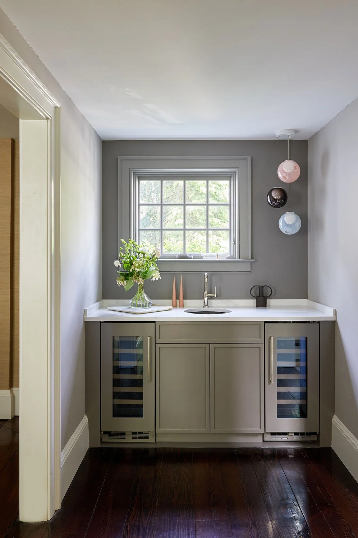 Full service wet bar with dual wine fridges, taupe painted cabinetry, quartz countertop, and sphere pendants in a Darien CT home by Ginger Lemon Indigo