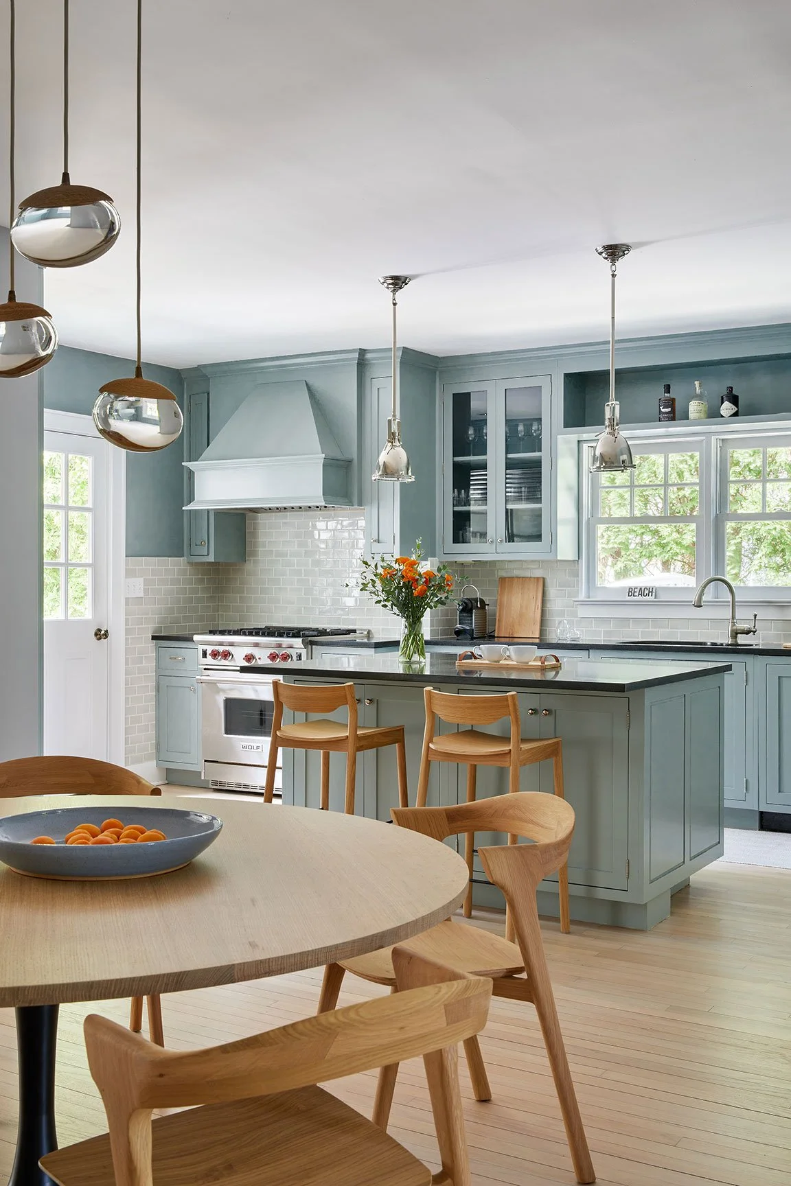 Duck egg blue kitchen with round oak dining table, Wolf range, pebble glass pendants, and wood bar stools in a Southampton NY coastal home by Ginger Lemon Indigo