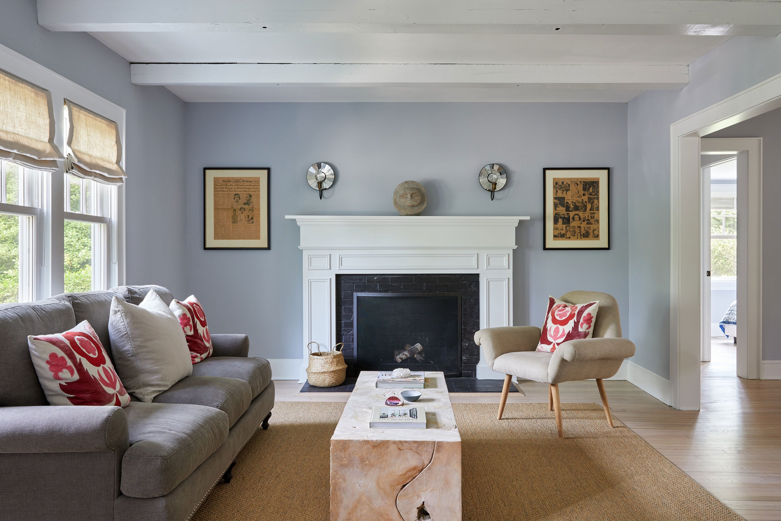 Living room with grey sofa, red floral pillows, mid century armchair, marble coffee table, and vintage sun face on white fireplace mantle in a Southampton NY home by Ginger Lemon Indigo