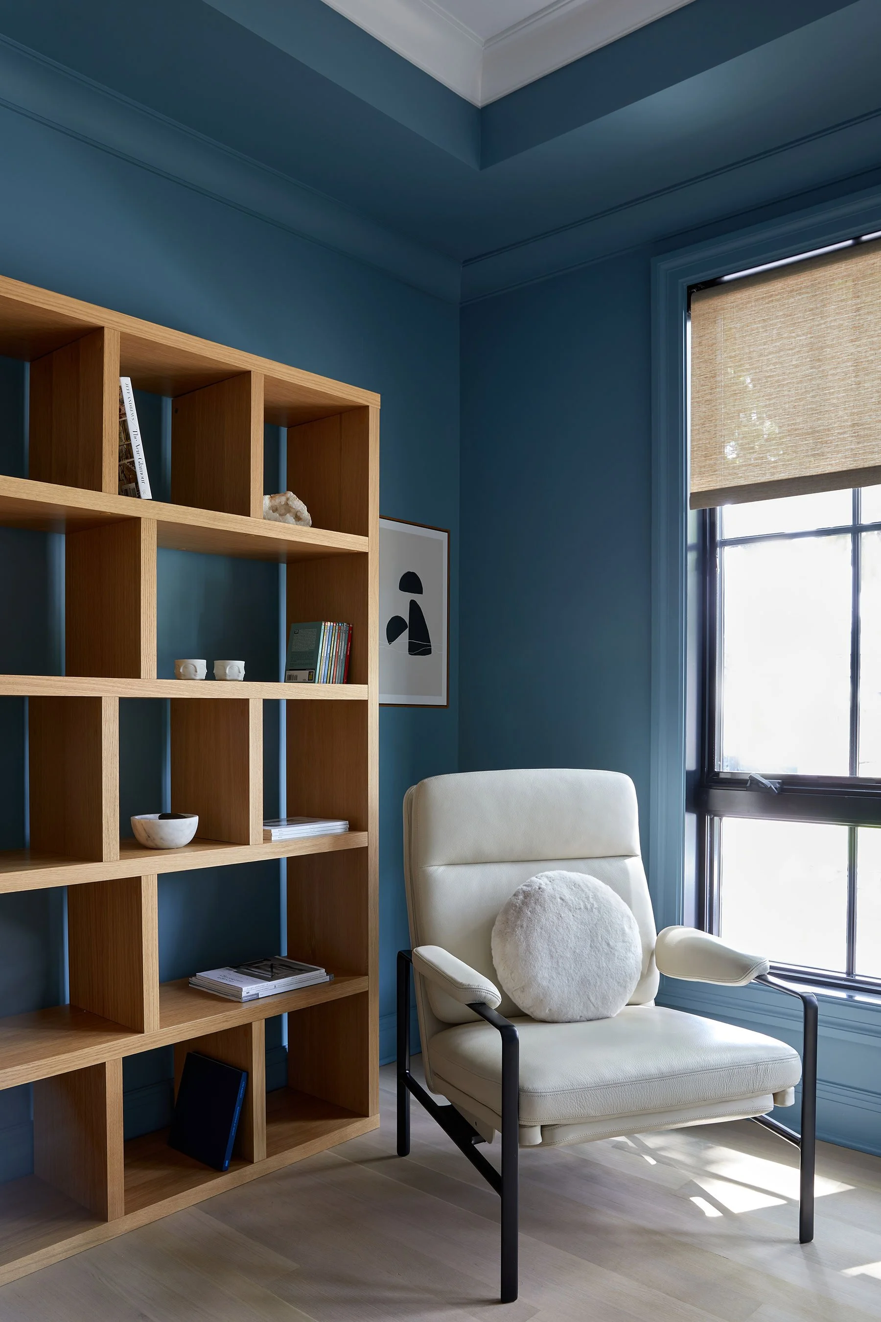 Home study with deep blue walls and ceiling tray, natural oak shelving, cream leather recliner, and woven roman shade in a Rye NY condo by Ginger Lemon Indigo