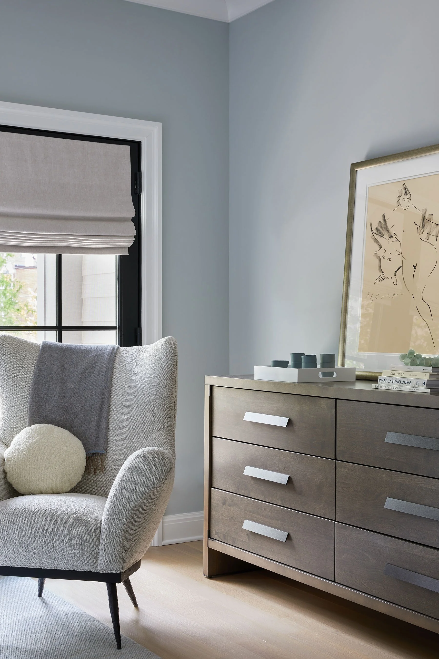 Bedroom reading corner with cream bouclé wingback chair, wood dresser, framed figurative artwork, and grey roman shade in a Rye NY condo by Ginger Lemon Indigo
