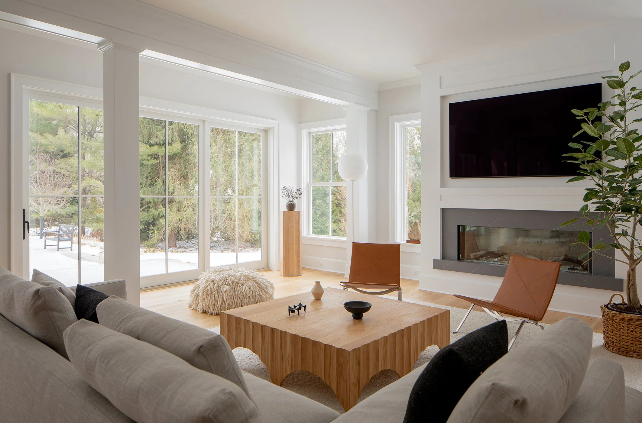 Nearby project in Westport CT family room with linen sectional, oak arched coffee table, pair of cognac leather PK22 chairs, linear fireplace and tall windows onto evergreens