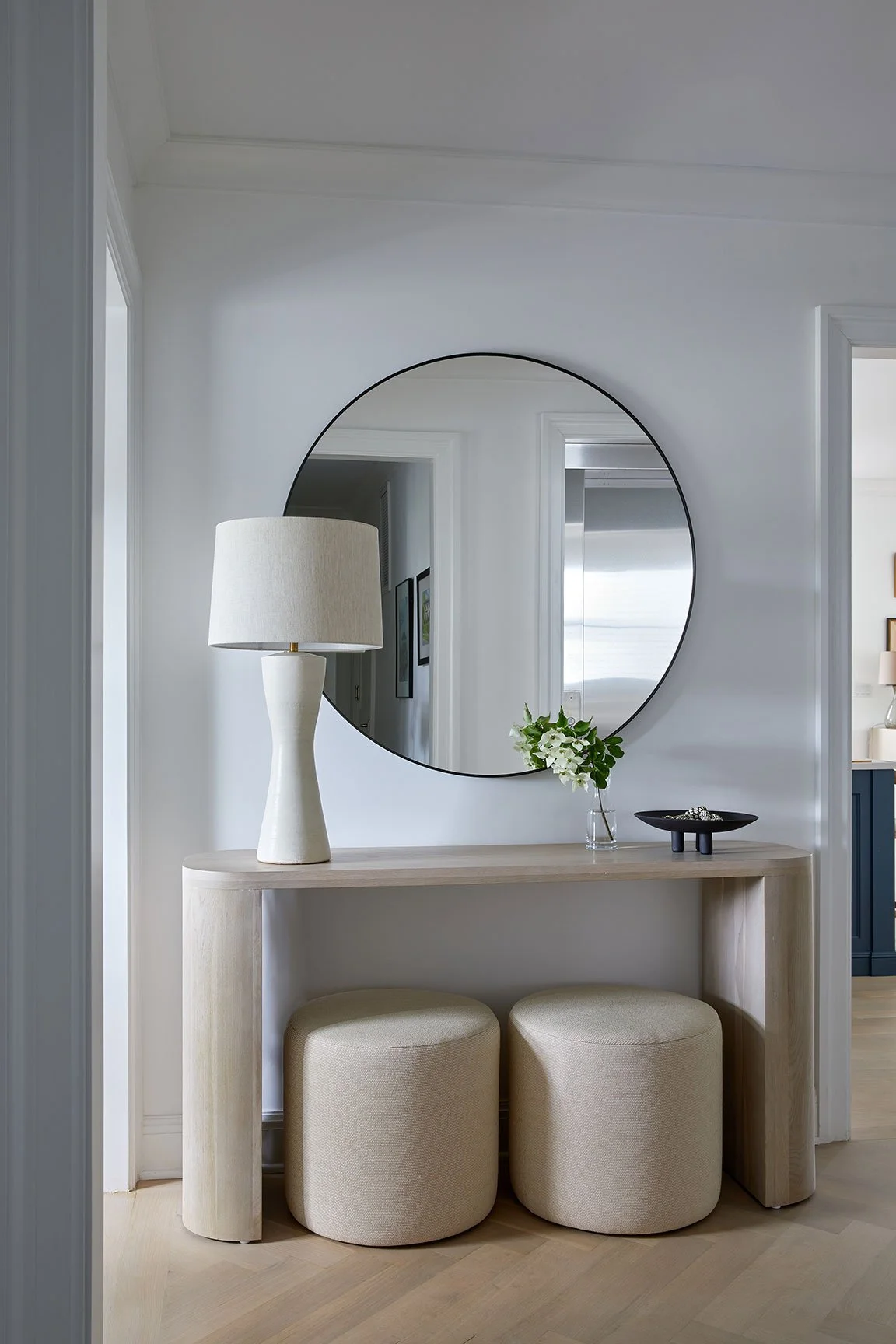 Modern entry with large round mirror, natural wood console table, ceramic table lamp, and two upholstered stools in a Rye NY condo by Ginger Lemon Indigo