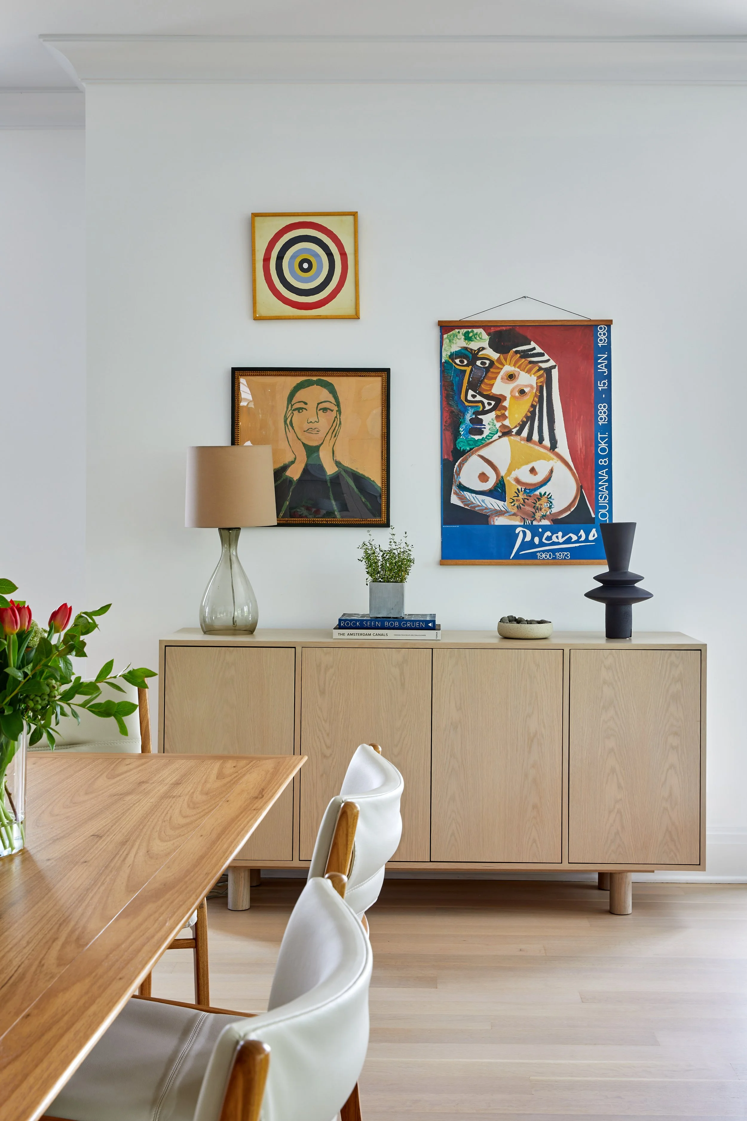 Dining area sideboard with natural oak credenza, Picasso exhibition poster, portrait painting, and glass table lamp in a Rye NY condo by Ginger Lemon Indigo