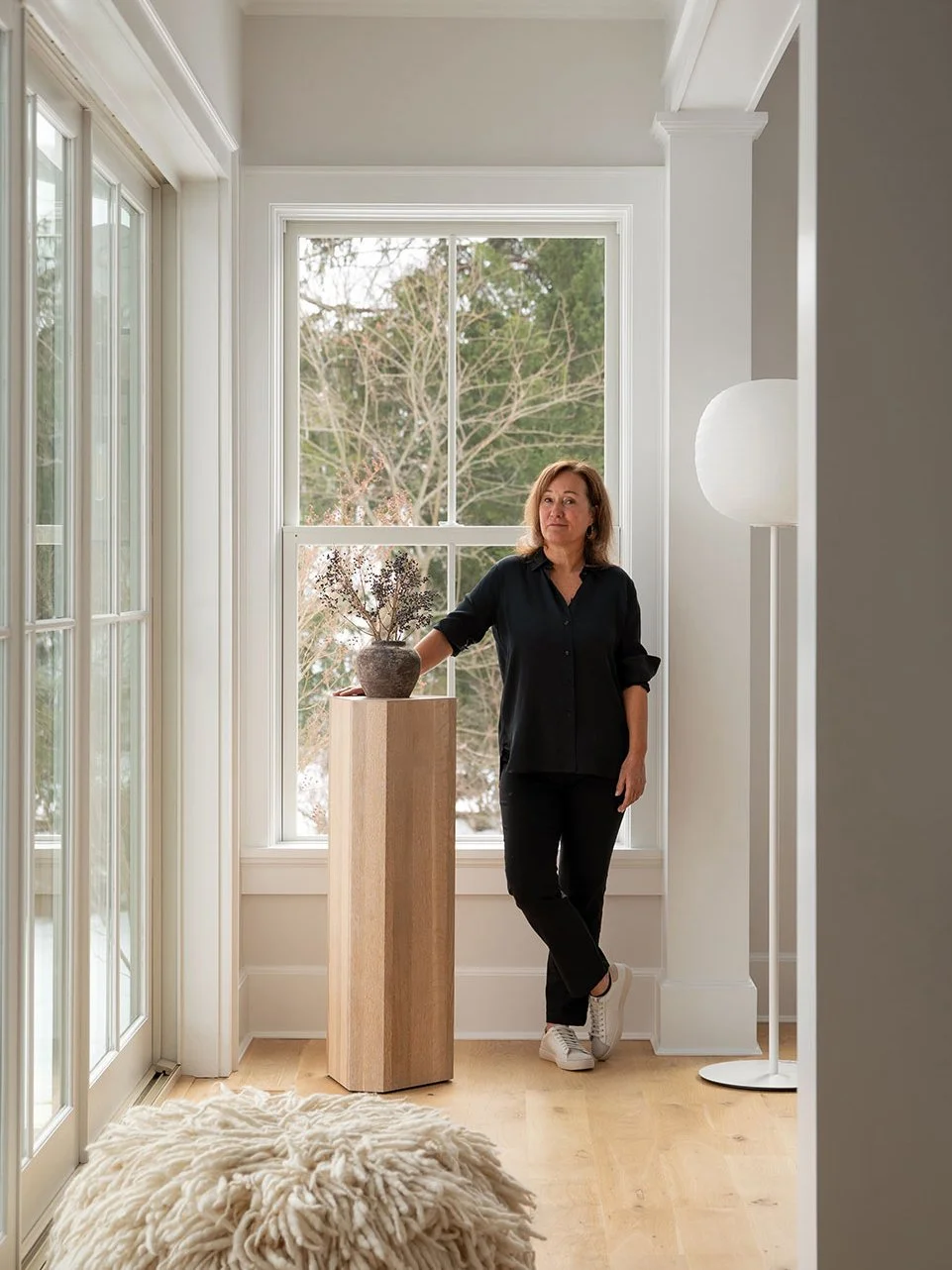 Barbra Fordyce interior designer standing in a light-filled Westport CT project space beside a ceramic vessel on a wood pedestal