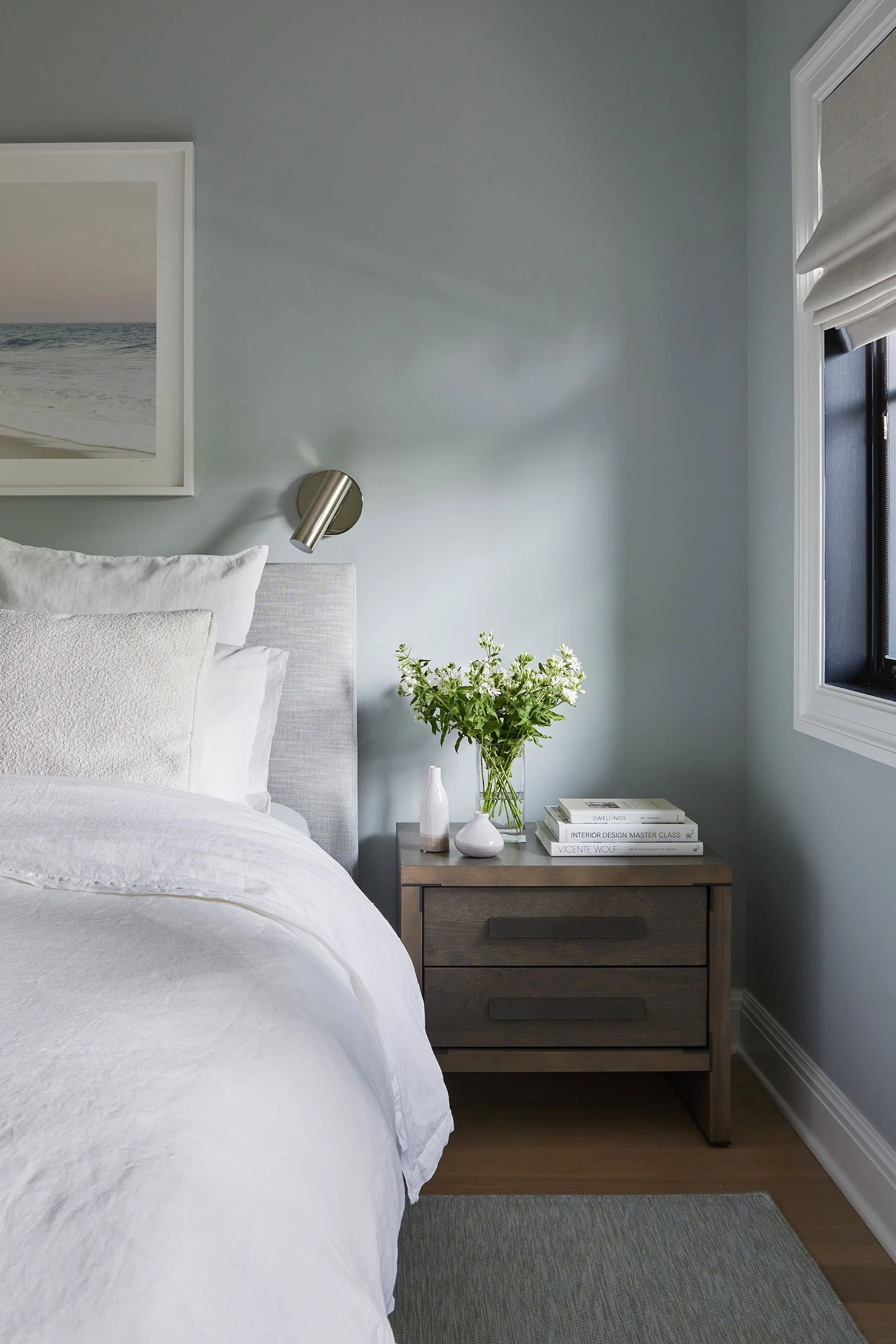 Primary bedroom detail with seafoam walls, upholstered headboard, wood nightstand, wall sconce, and fresh flowers in a Rye NY condo by Ginger Lemon Indigo