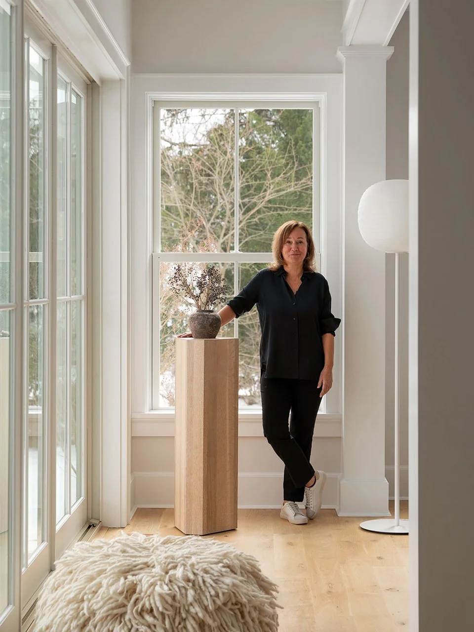 Barbra Fordyce interior designer standing in a light-filled Westport CT project space beside a ceramic vessel on a wood pedestal