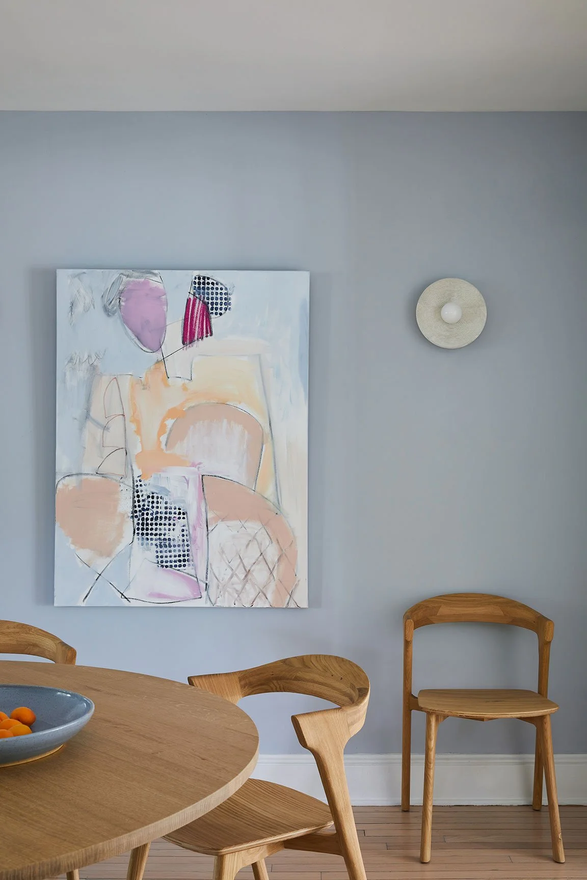 Breakfast area detail with custom round oak table on black pedestal, Allison Meyler painting on coastal blue wall, and organic disc wall sconce in a Southampton NY home by Ginger Lemon Indigo