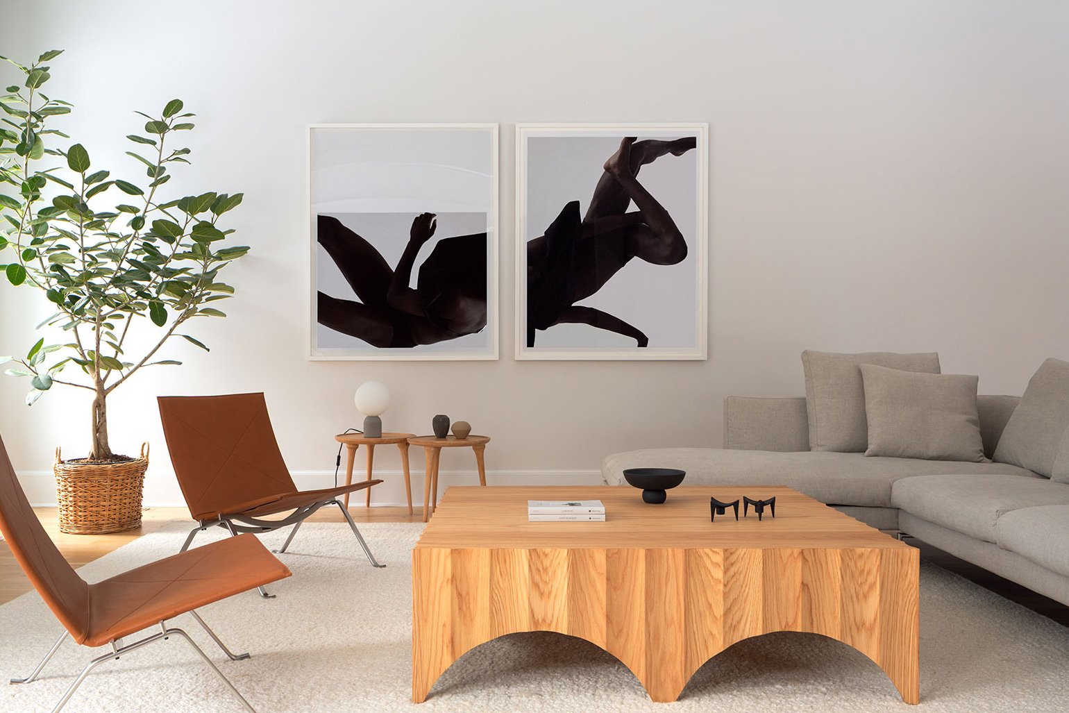 Living room with a large gray sectional sofa, a wooden coffee table, and modern wall art of two abstract black silhouette figures. There's a tall potted plant on the left and a small side table with decor items.