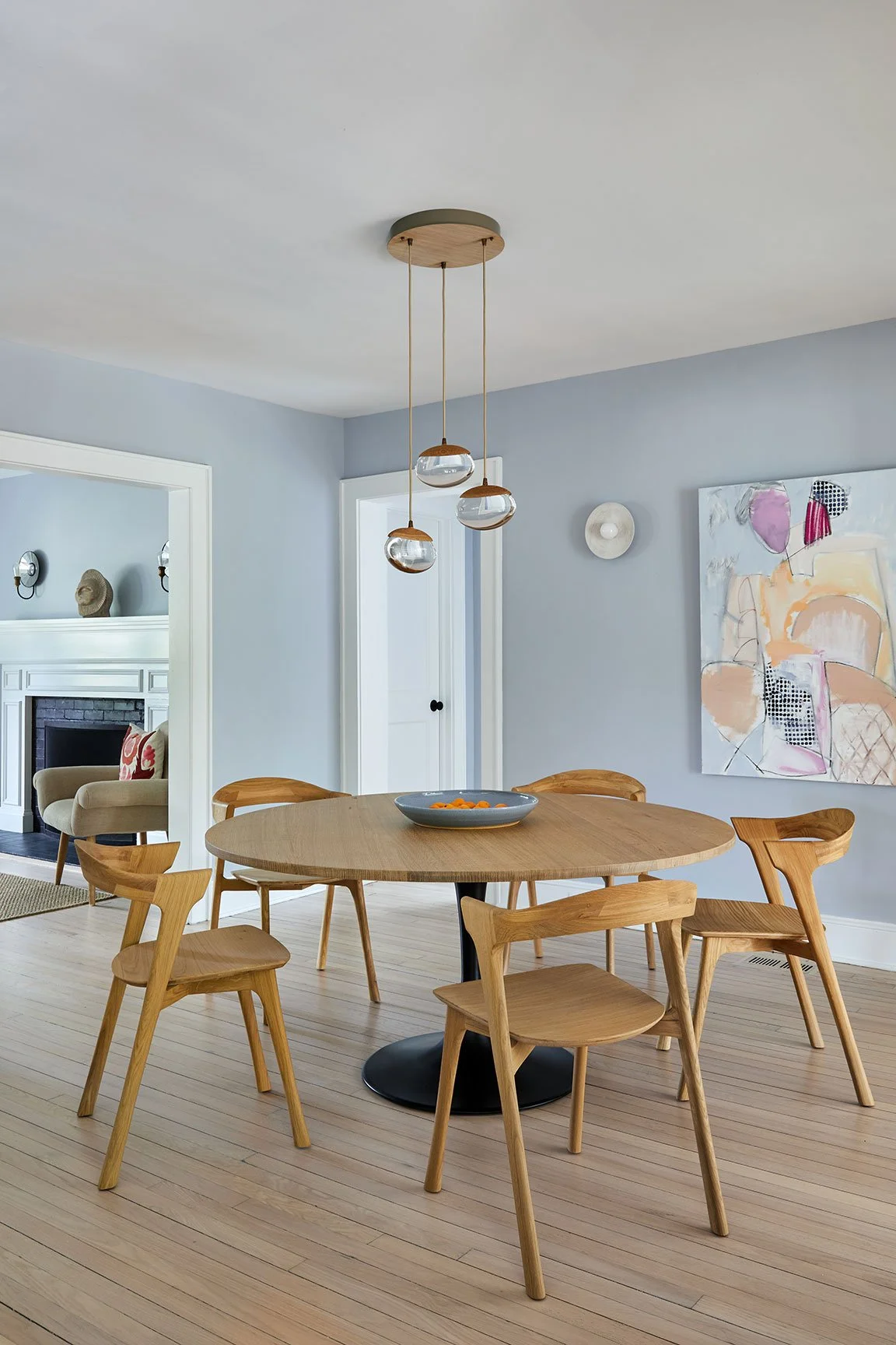 Custom round oak dining table on black metal pedestal, pebble glass pendant, and Allison Meyler painting in a Southampton NY coastal renovation by Ginger Lemon Indigo