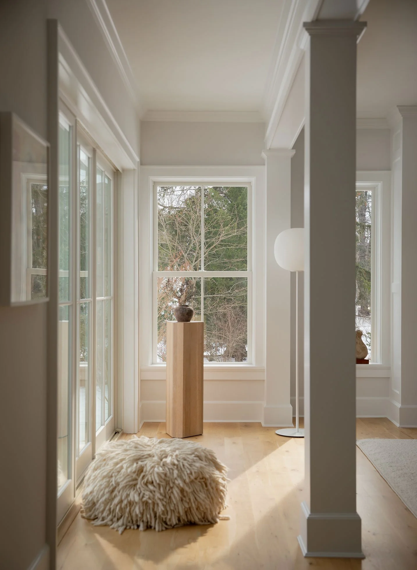 Nearby project in Westport CT sunlit hallway with oak pedestal and ceramic vessel, globe floor lamp, shaggy cream wool pouf and tall windows onto bare trees