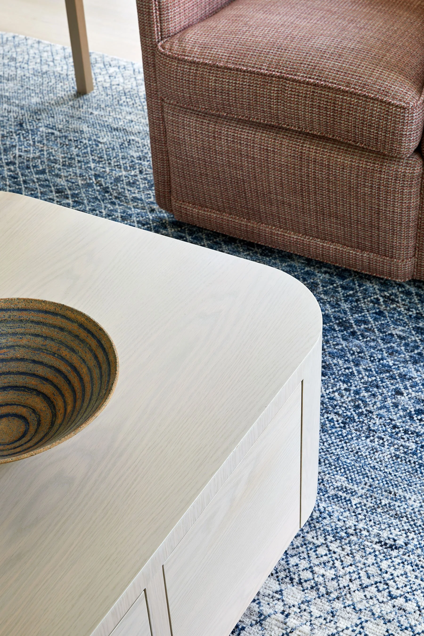 Detail of custom rounded coffee table with ceramic bowl, and woven upholstered chair on blue patterned rug in a Rye NY condo by Ginger Lemon Indigo