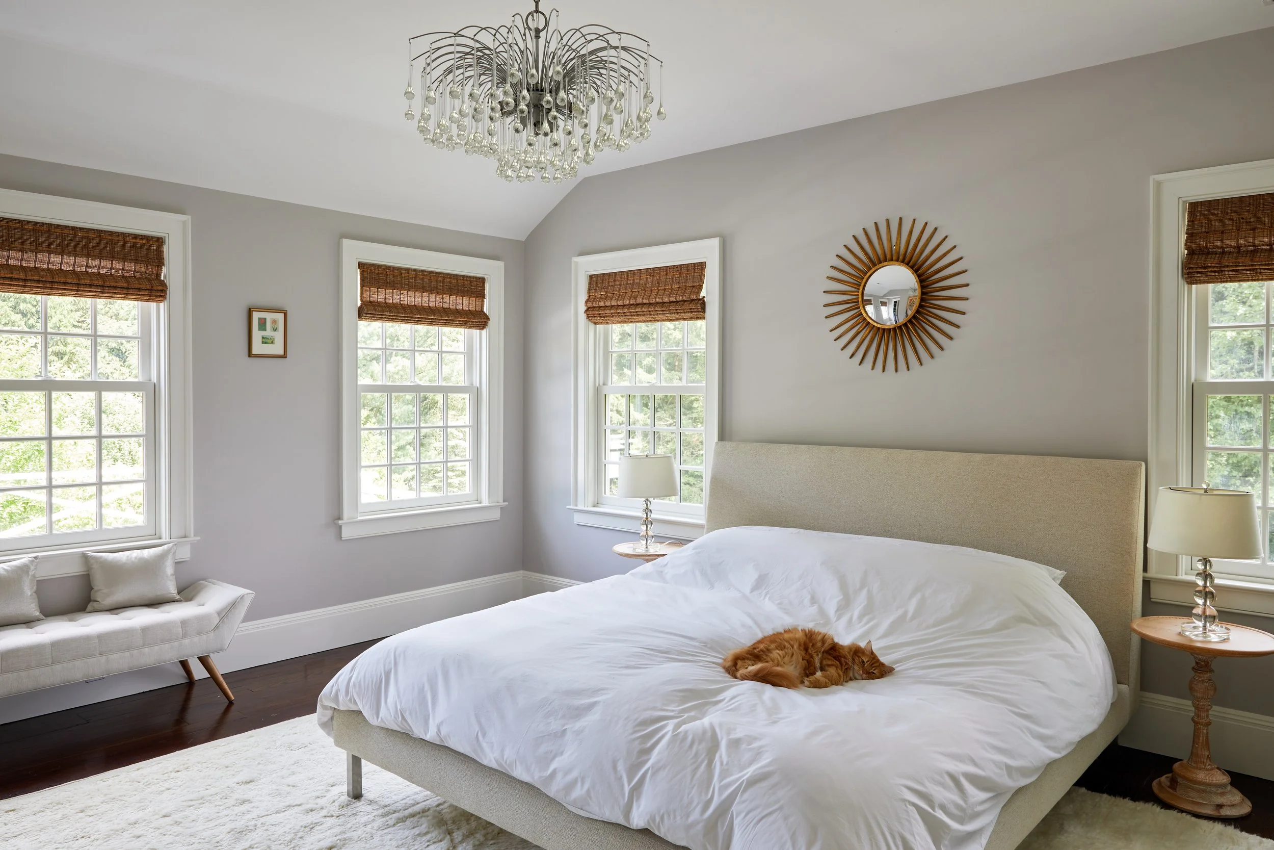 Nearby Darien CT bedroom with upholstered bed, crystal drop chandelier, gilt sunburst mirror, bamboo Roman shades and ginger cat on the duvet