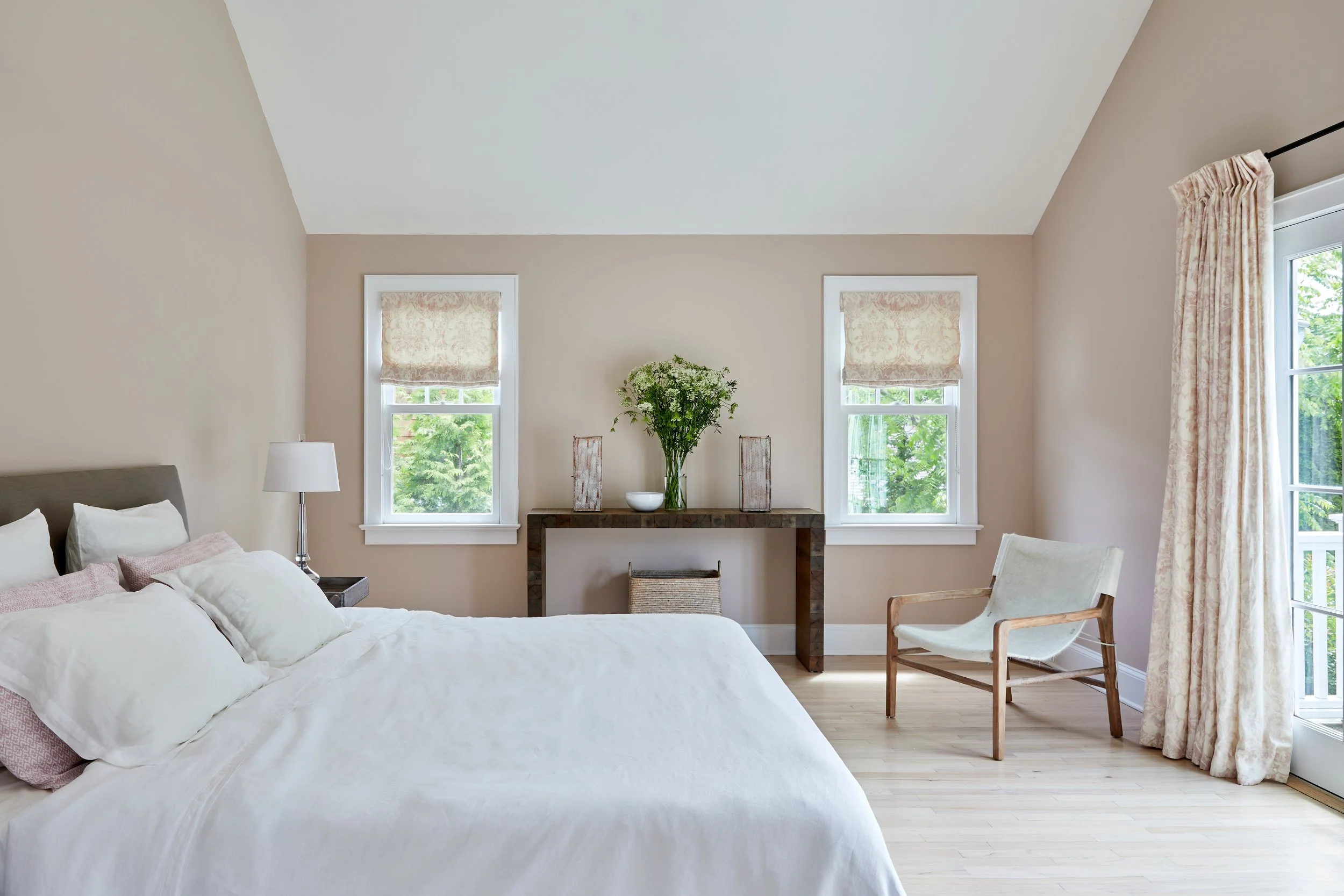 Primary bedroom in a Southampton NY shingle-style home by Ginger Lemon Indigo with warm taupe walls, white linen bedding, vaulted ceiling, vintage console and a sling lounge chair