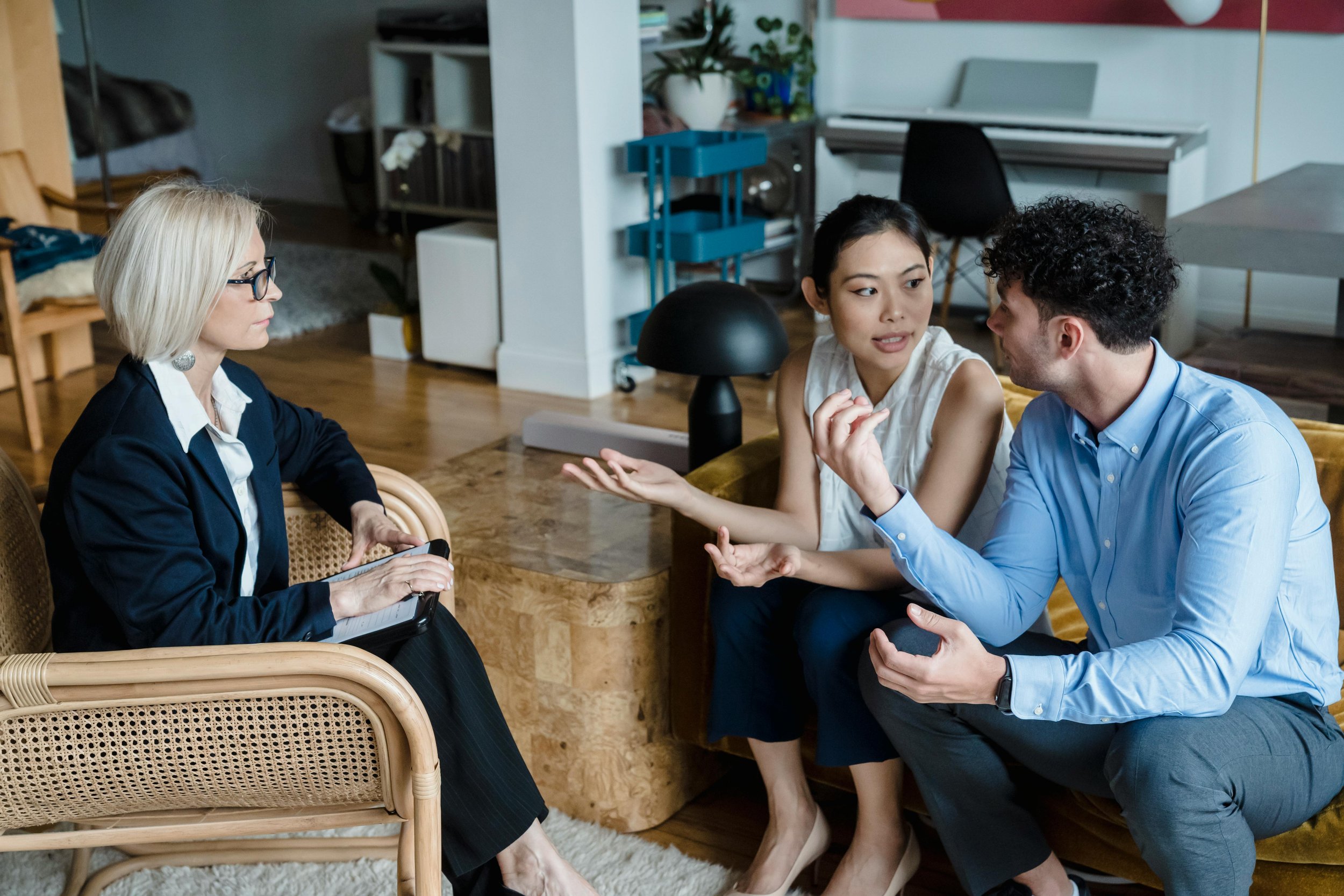 Three people having a discussion in a home or office setting. Two women and one man are seated around a table. The woman on the left has blonde hair, glasses, and is taking notes with a tablet. The woman in the middle and the man are engaged in a conversation, with the woman gesturing as she talks.