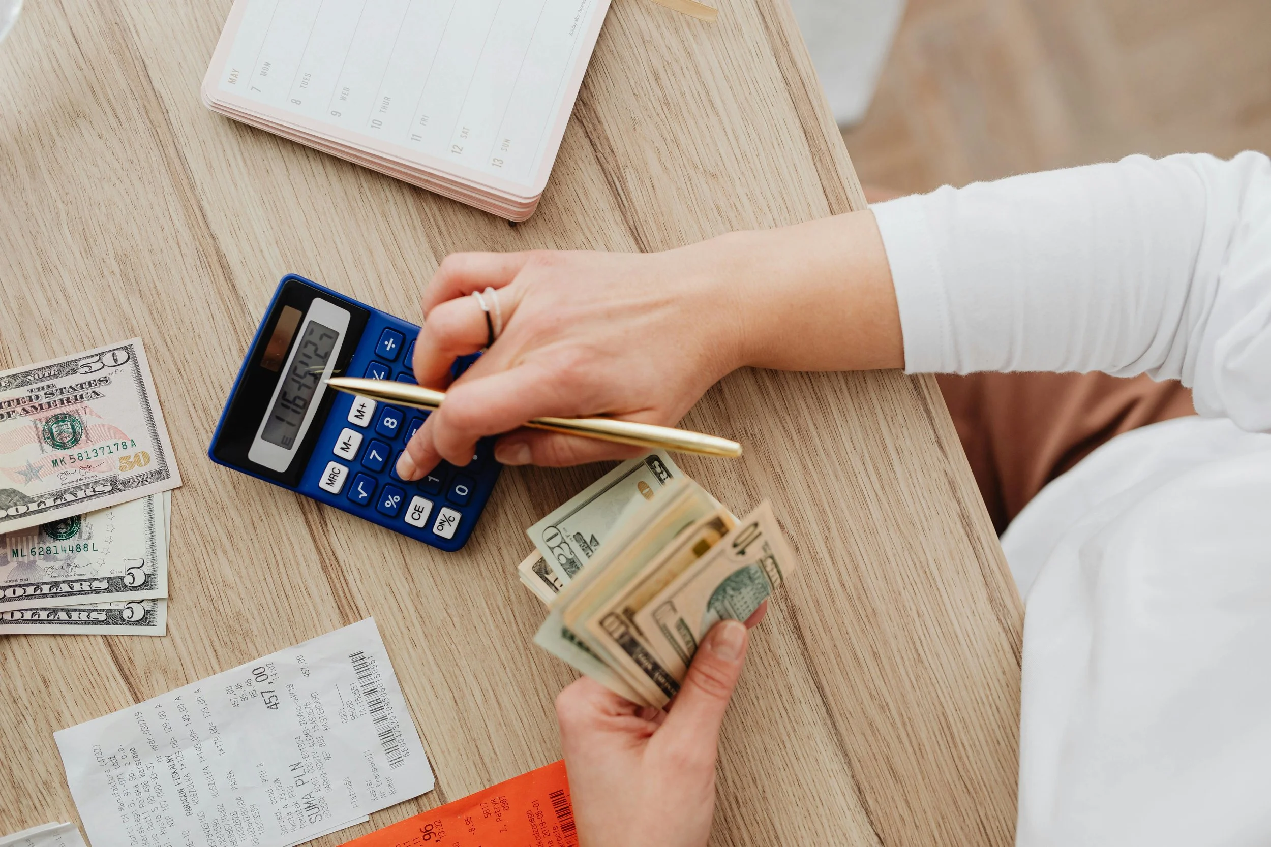 Person counting cash with a calculator on a wooden table, surrounded by receipts and money.