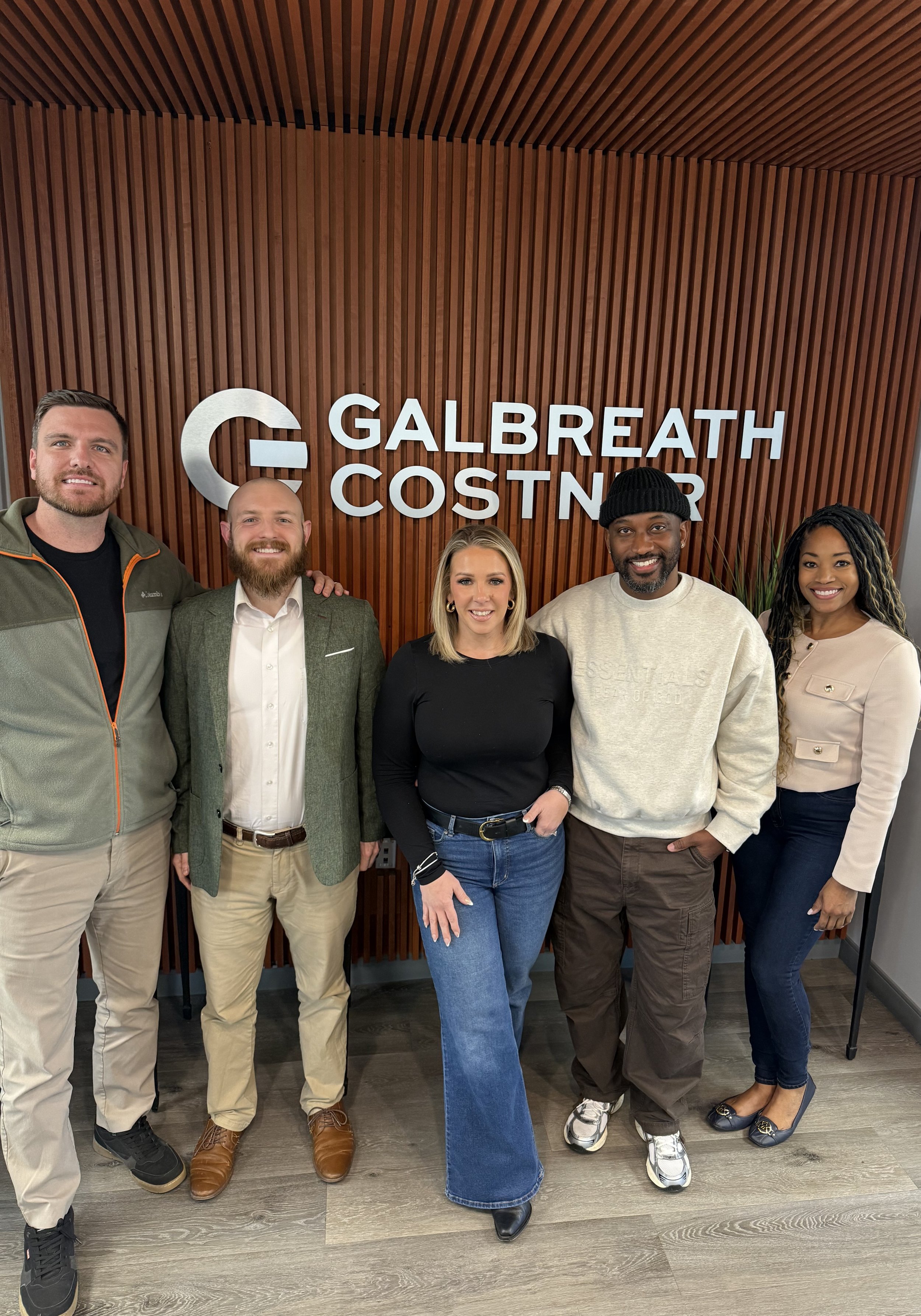 Group of six diverse professionals standing in front of a wood-paneled wall with a company sign that says 'Galbreath Construction.'
