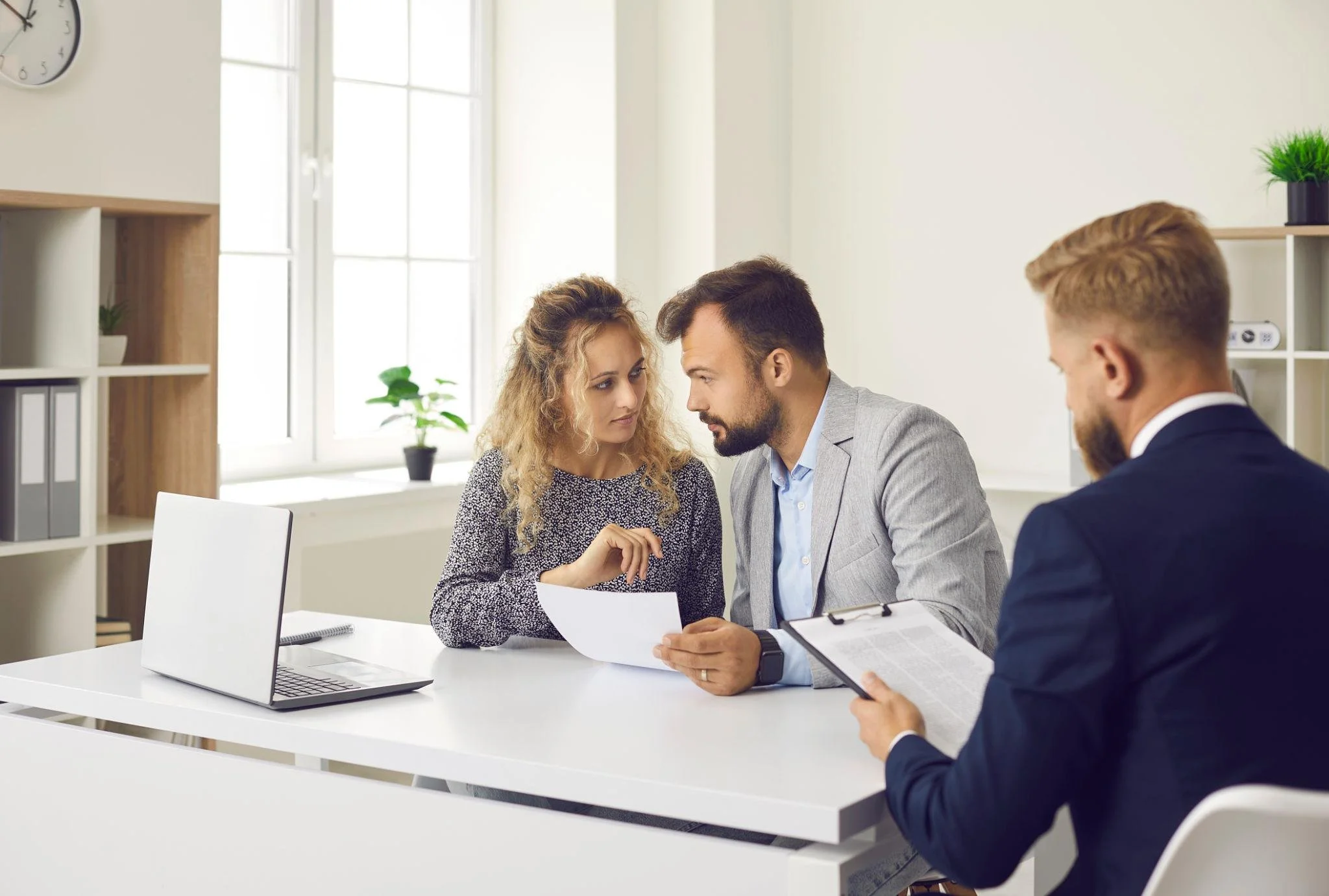 Three business professionals having a discussion in an office, with two sitting at a white table and one standing, looking at a document.