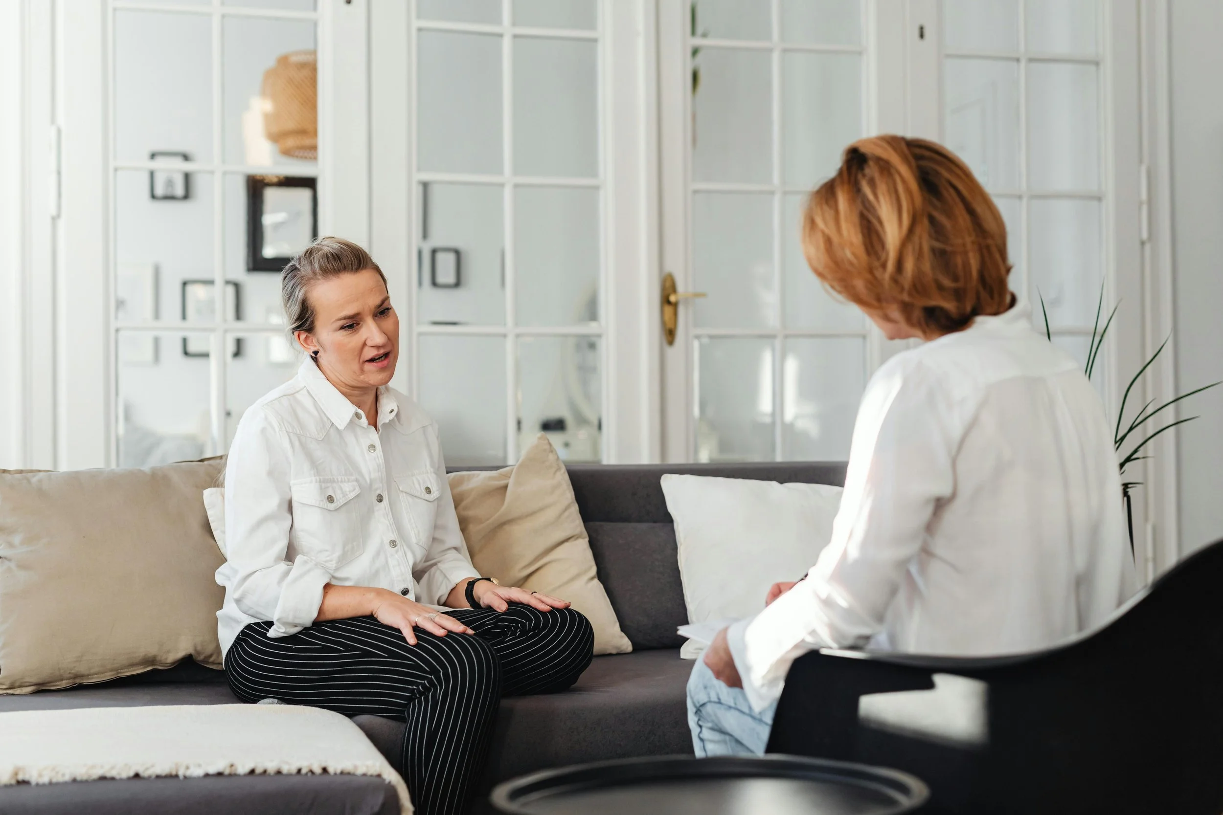 Two women having a conversation in a living room. One woman is sitting on a couch and the other is standing in front of her, with the background featuring a white wall with shelves and decorative items.