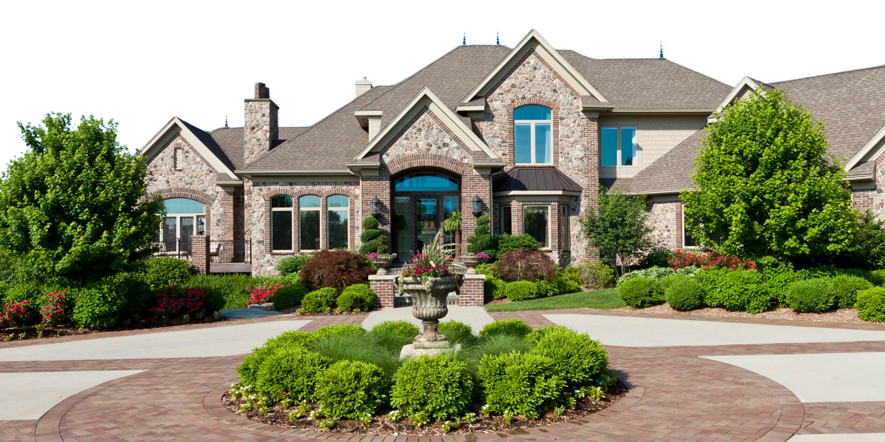 A large, elegant brick house with multiple gables, surrounded by well-maintained green landscaping and trees, with a decorative stone fountain in the front yard.