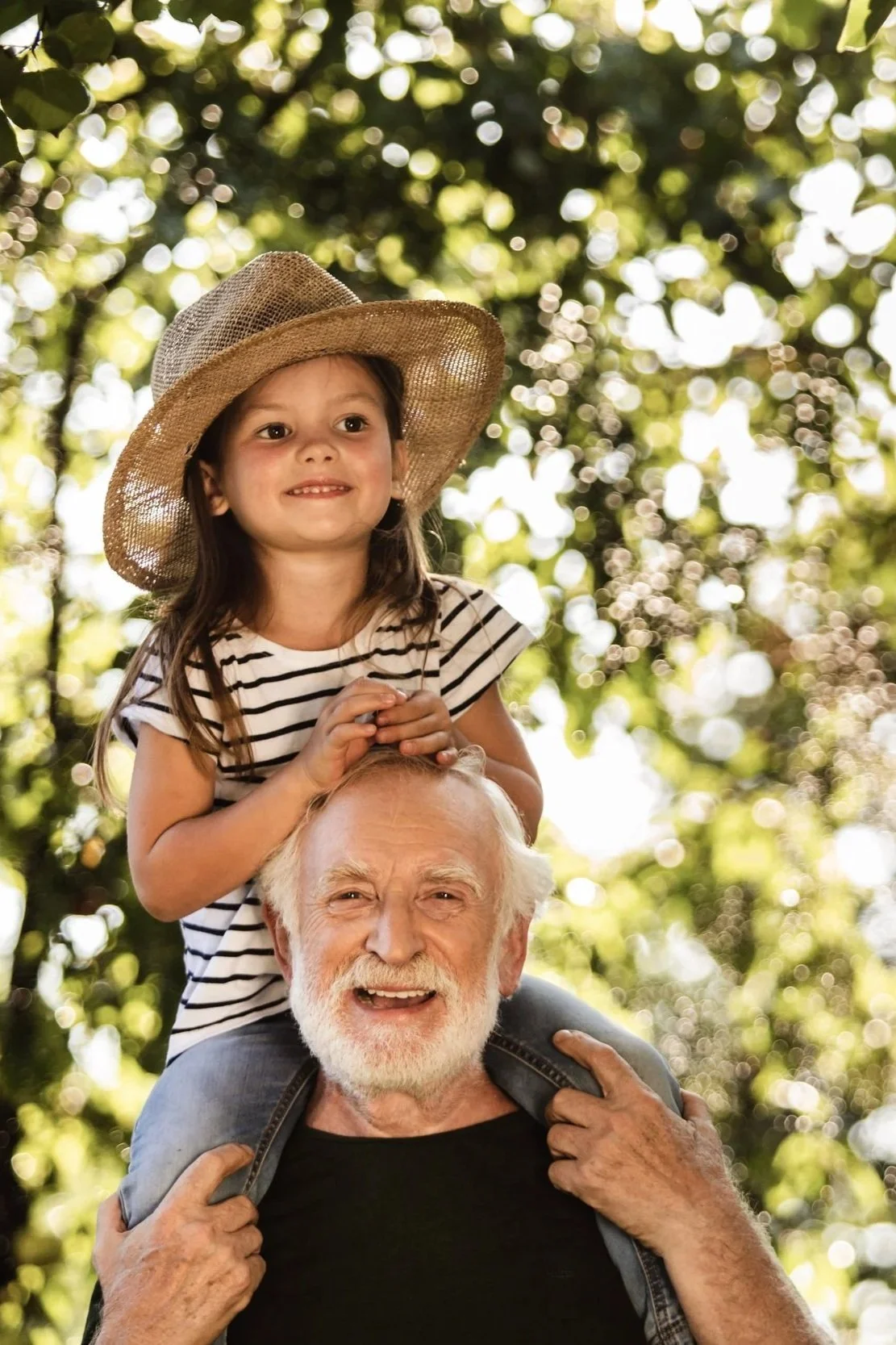 A young girl with long dark hair, wearing a striped shirt and straw hat, sitting on an older man's shoulders in a park with green trees and sunlight in the background.