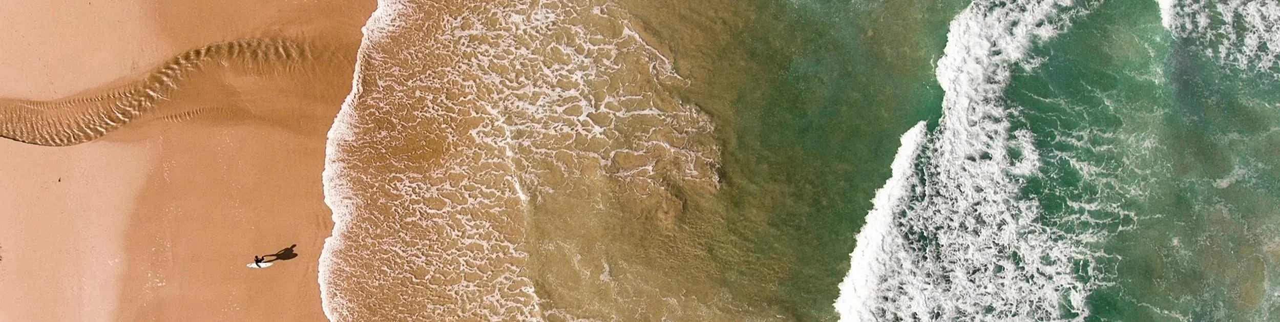 An aerial view of a sandy beach with a surfer carrying a surfboard, and ocean waves crashing onto the shore.