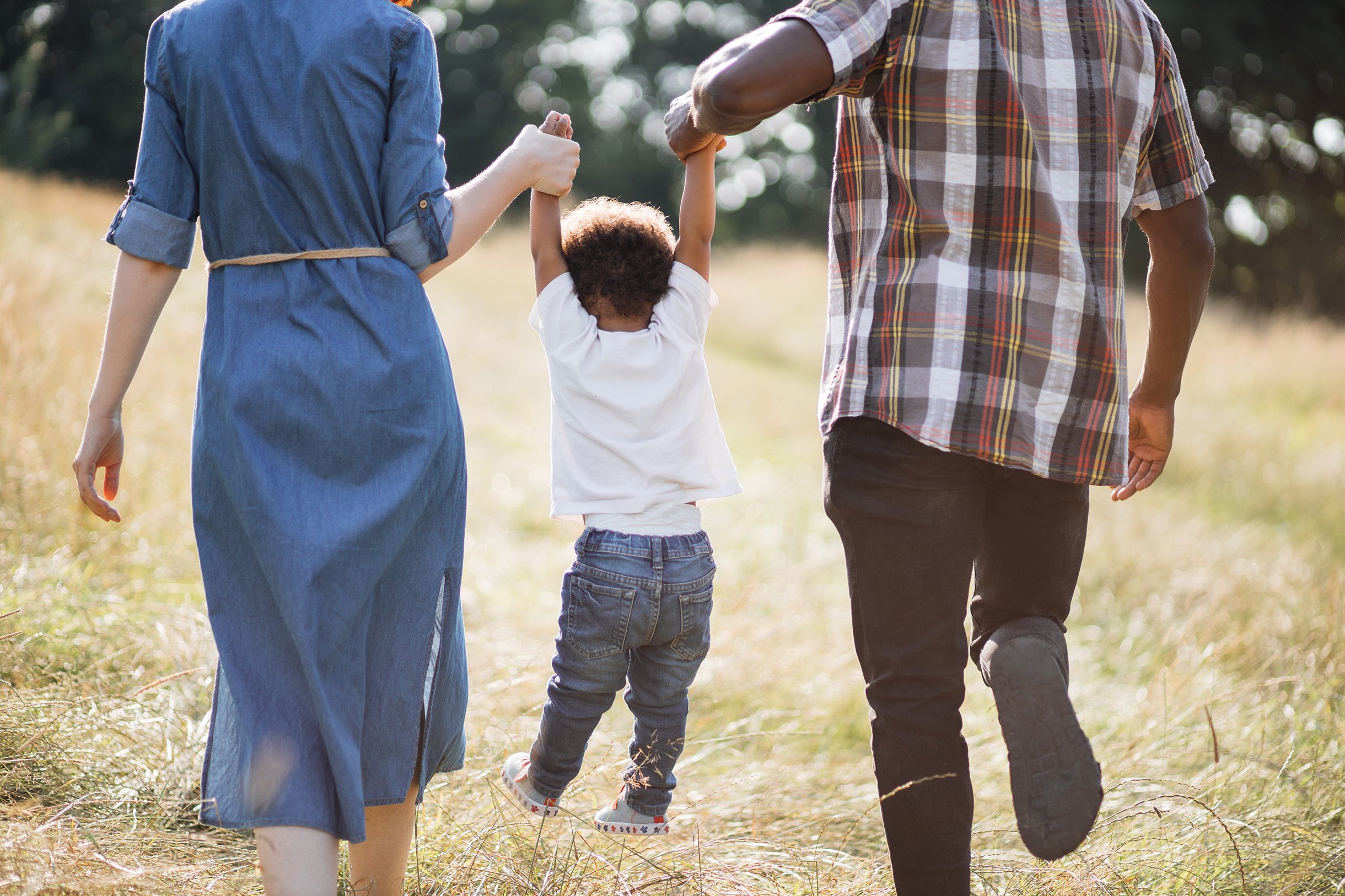 A young girl is walking outdoors with two adults, holding their hands. They are walking on a grassy field with trees in the background. The girl is wearing a white shirt, jeans, and sneakers with floral patterns. The adults are dressed casually, one in a denim dress and the other in a plaid shirt and dark pants.