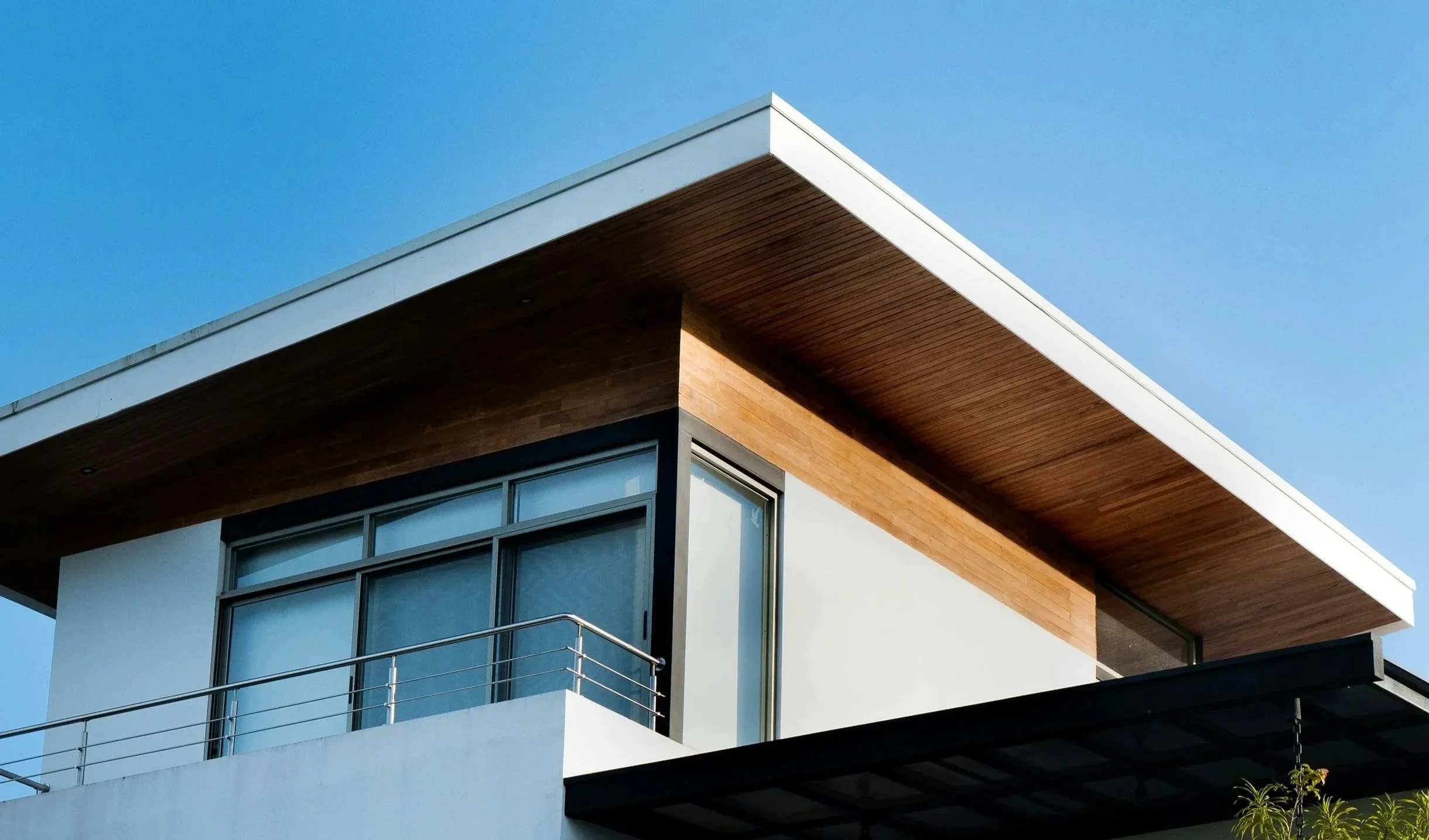 Close-up of a modern building's corner with white walls, large glass windows, wooden accents, a metal railing, and a clear blue sky in the background.