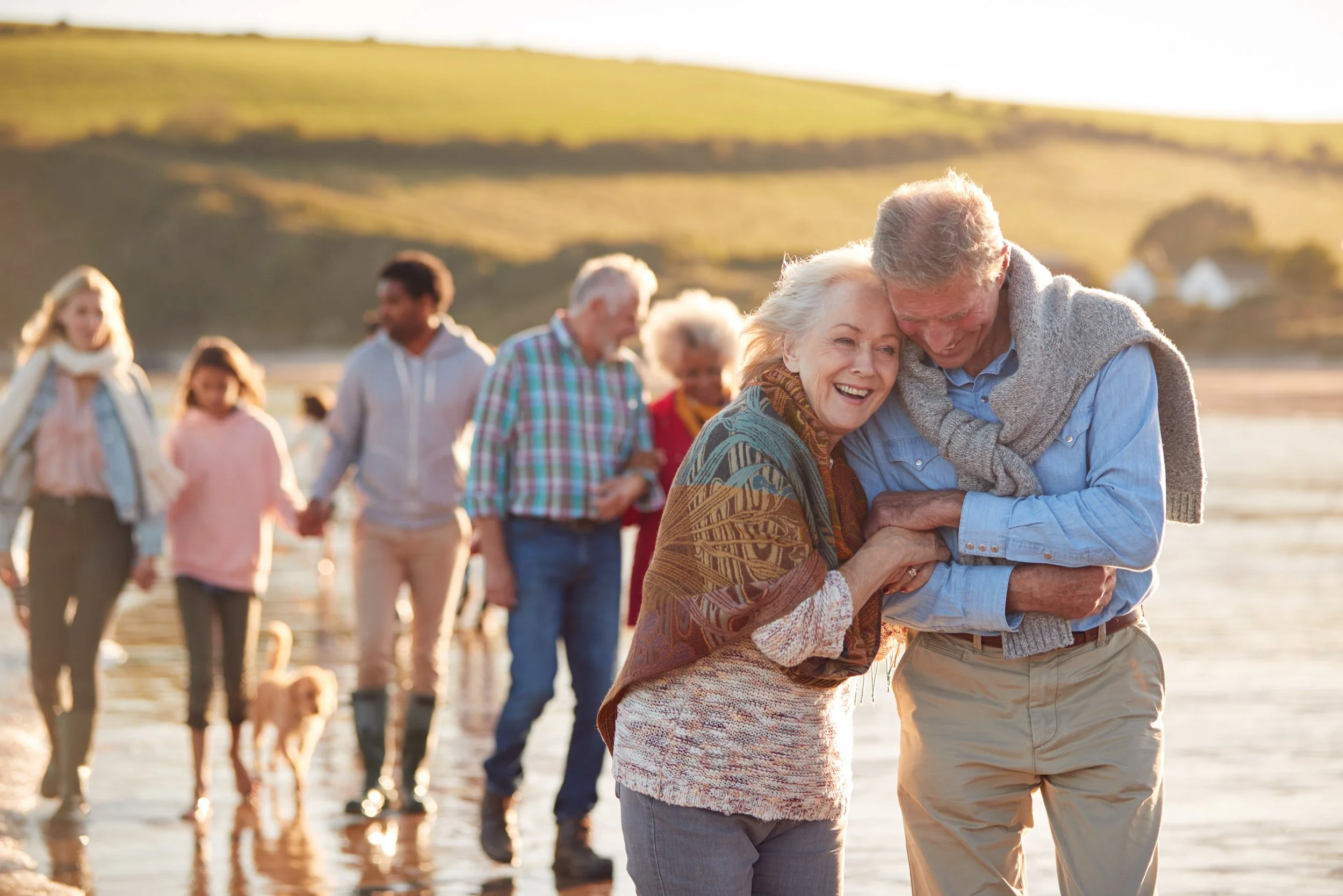 An elderly couple smiling and hugging on a beach with a group of people walking behind them during sunset.