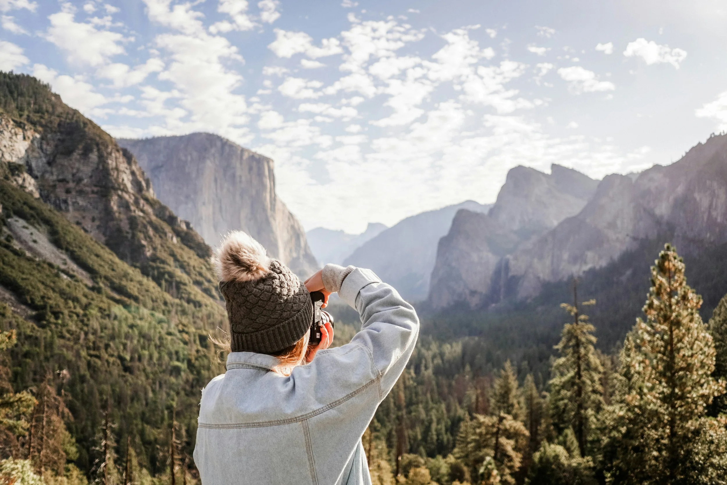 Child wearing a knit hat and gray hoodie taking a photograph with a camera of the scenic mountainous landscape with cliffs and forested areas under a partly cloudy sky.