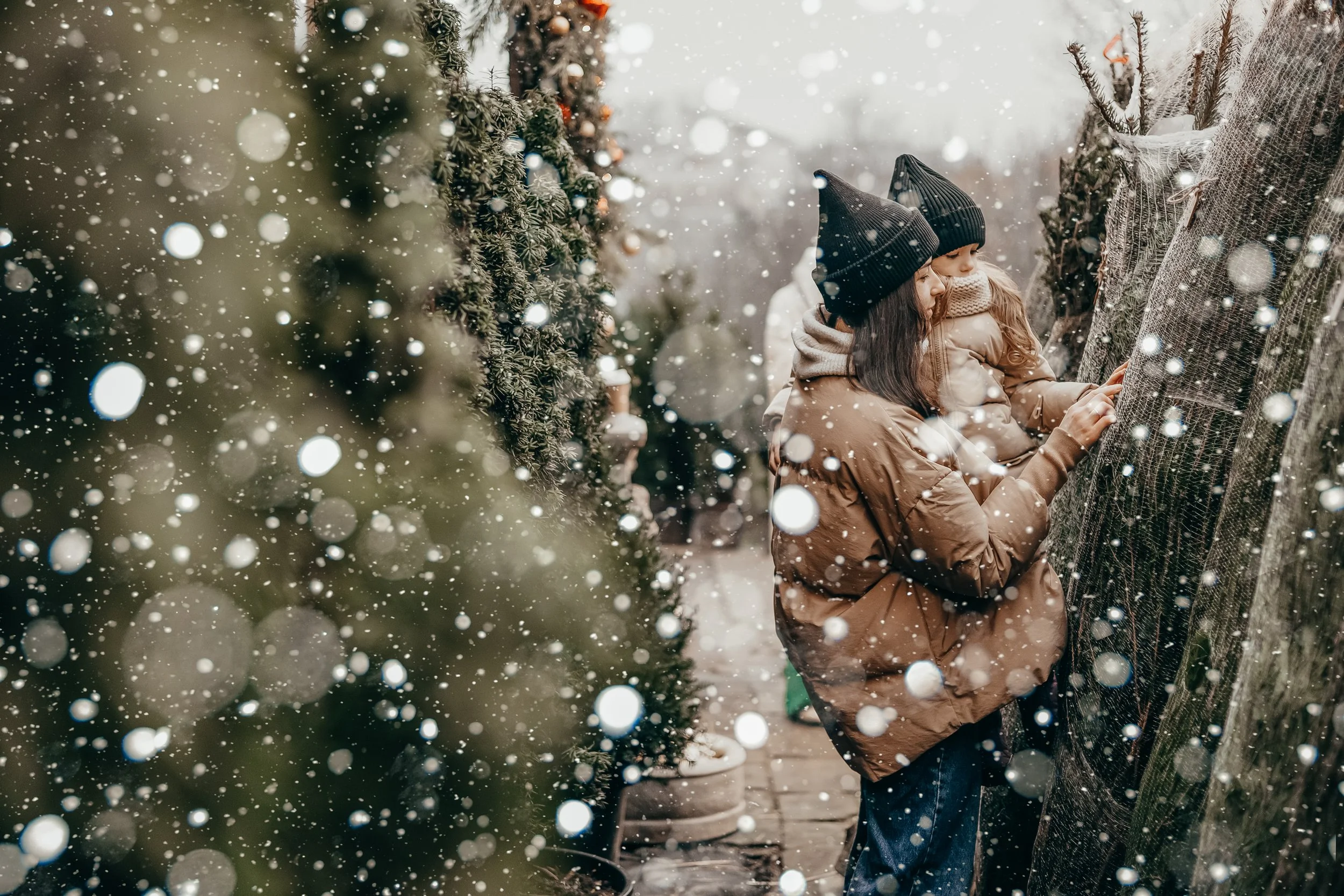 Two children in winter clothing touching a Christmas tree outdoors in snowfall.