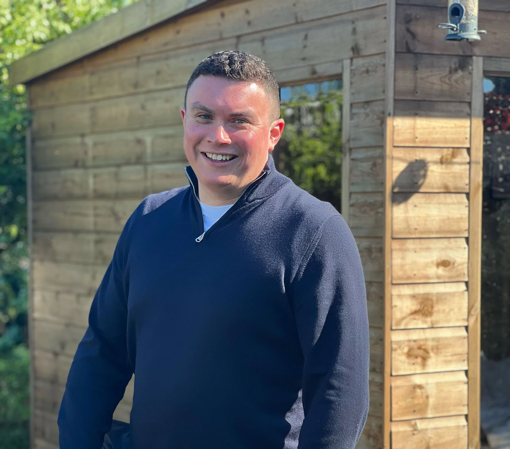 A smiling man in a navy blue zip-up jacket standing outdoors with a wooden shed in the background under a green leafy tree.