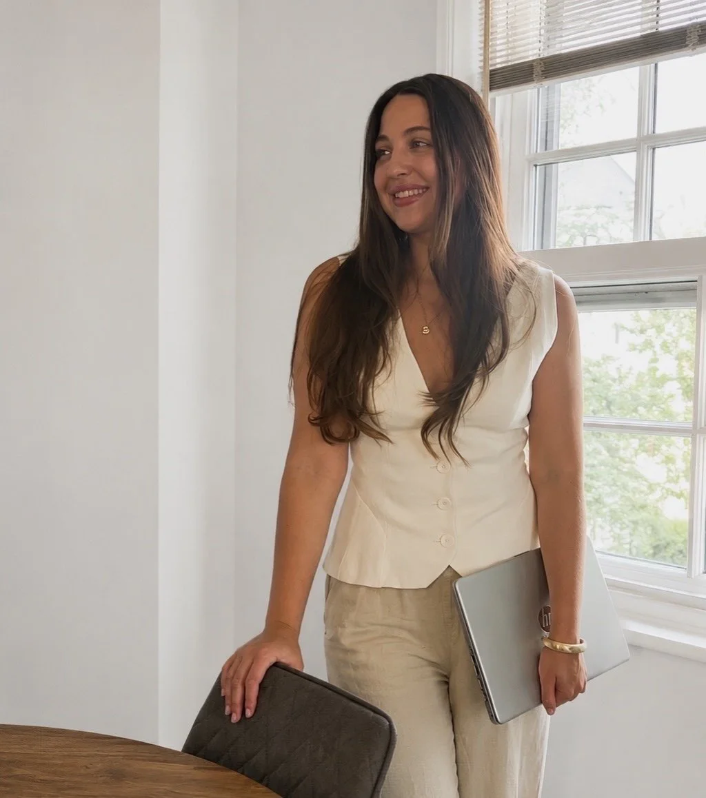 A woman with long brown hair, wearing a cream sleeveless blouse and beige pants, smiling while holding a closed laptop and standing by a table in a bright room with large windows.