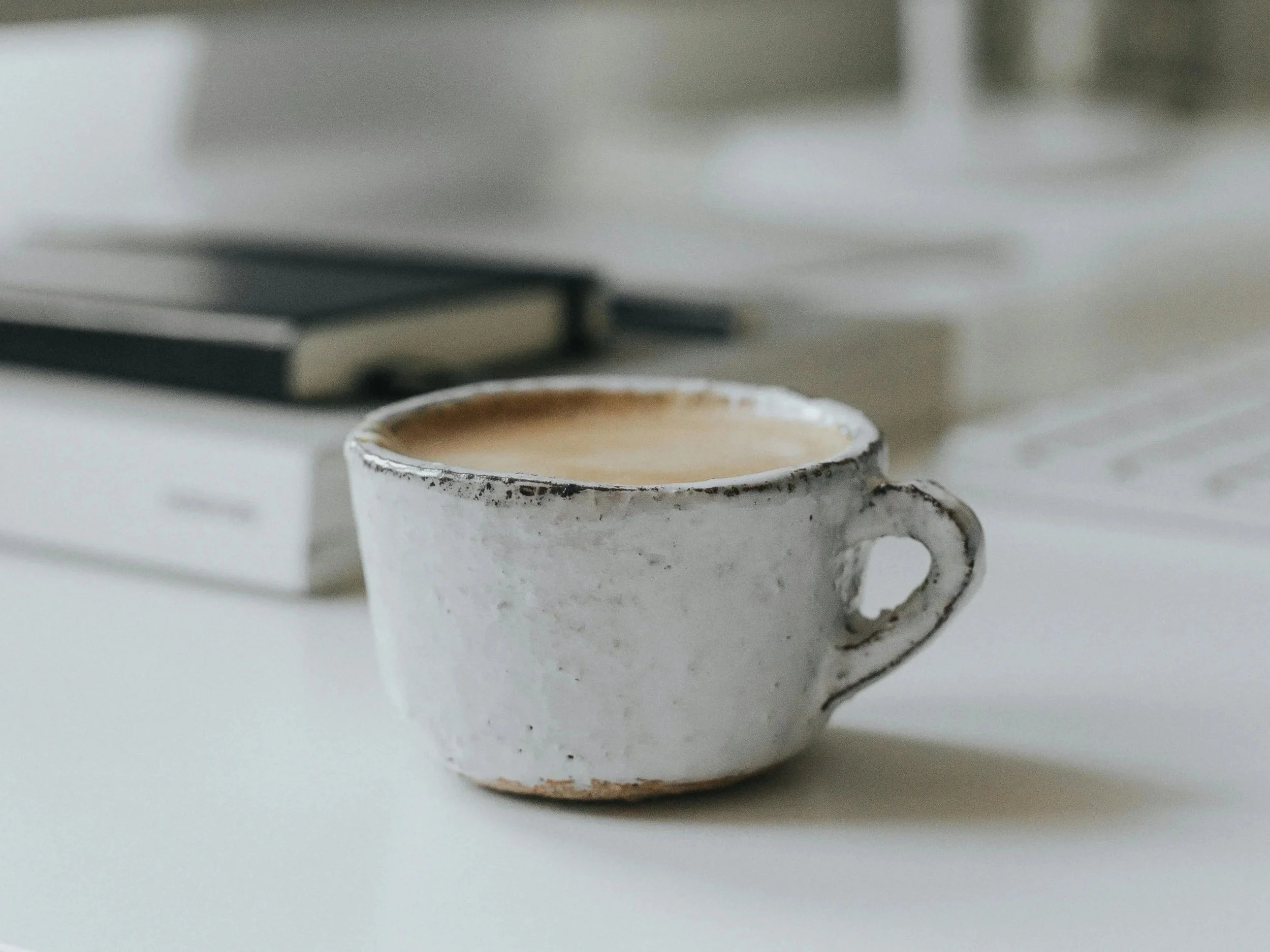 Close-up of a white ceramic mug with a textured finish, filled with coffee, on a white surface with a blurred background showing a closed book and part of a laptop.