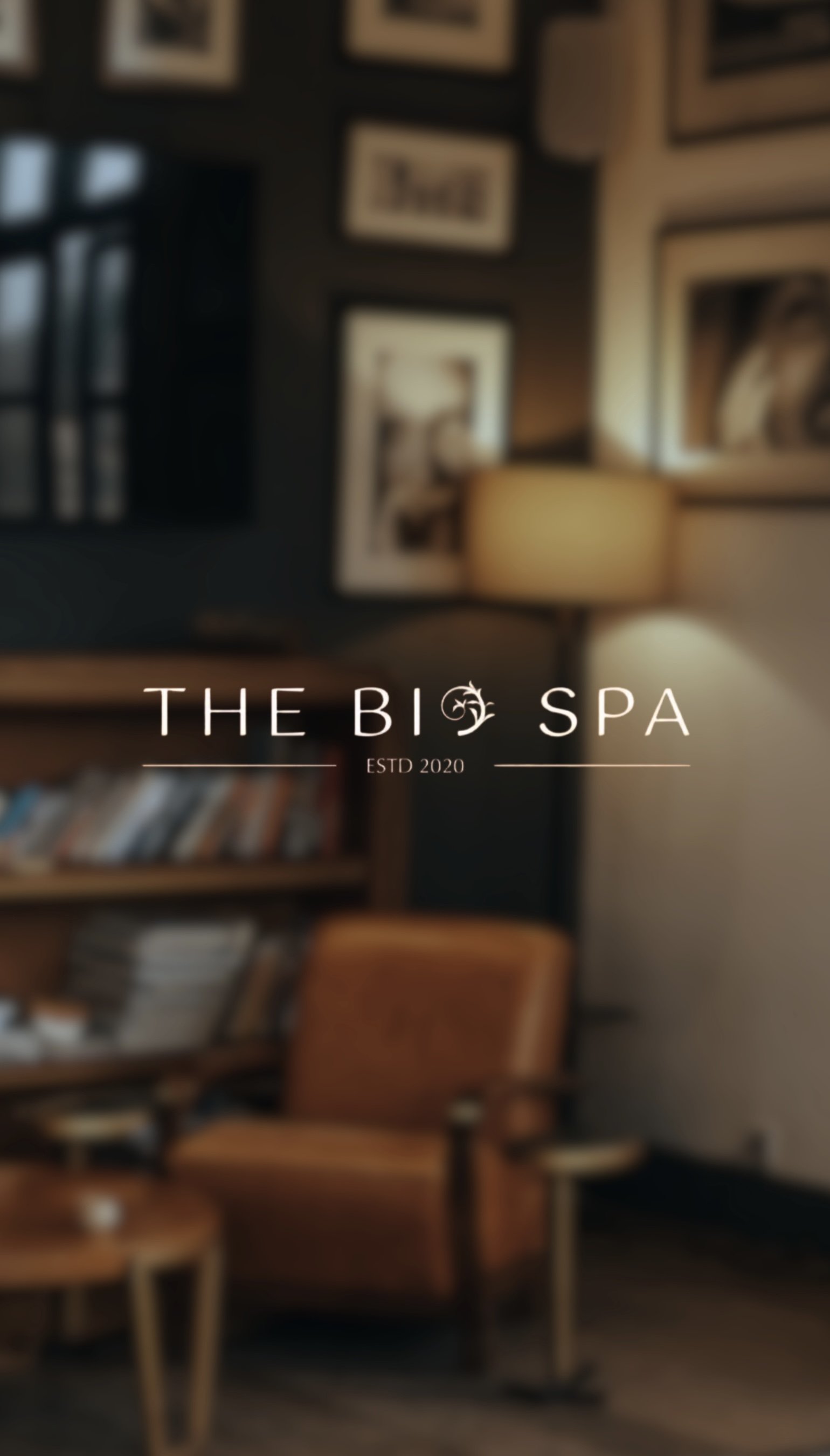 Blurred image of a cozy room with a bookshelf, framed pictures on the wall, a lamp, and a wooden chair. Text overlay reads 'The Big Spa ESTD 2020' as a logo of hotel for branding project