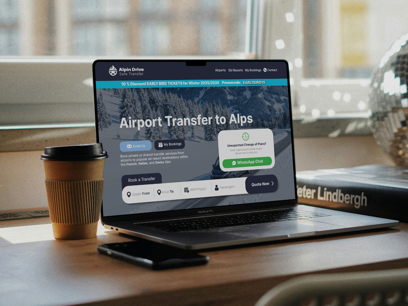 Laptop on a desk displaying a website for airport transfer to Alps, with a coffee cup and smartphone nearby, in an indoor setting with a window and a book in the background.