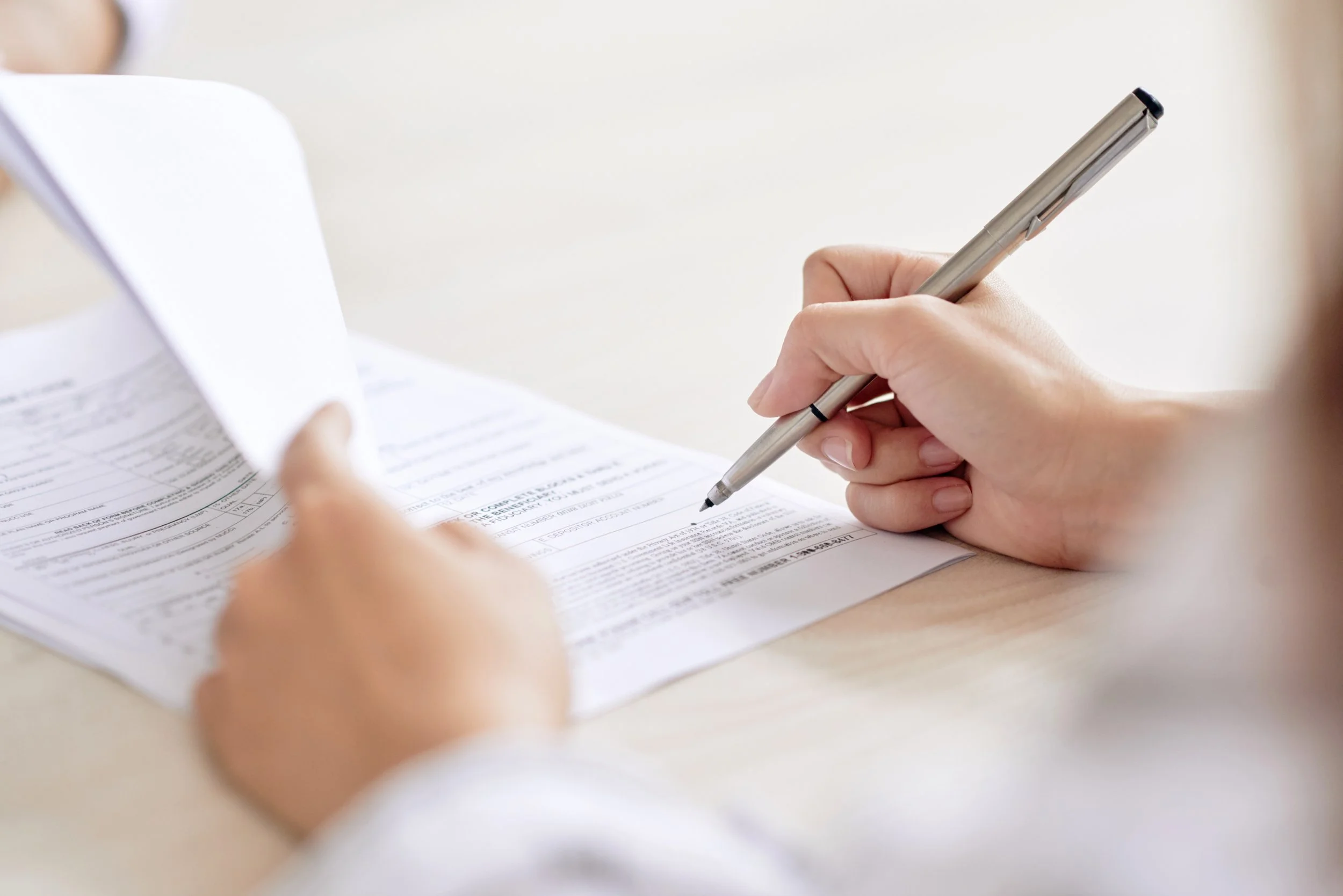 A person is holding a silver pen and filling out a form or document on a light-colored desk.