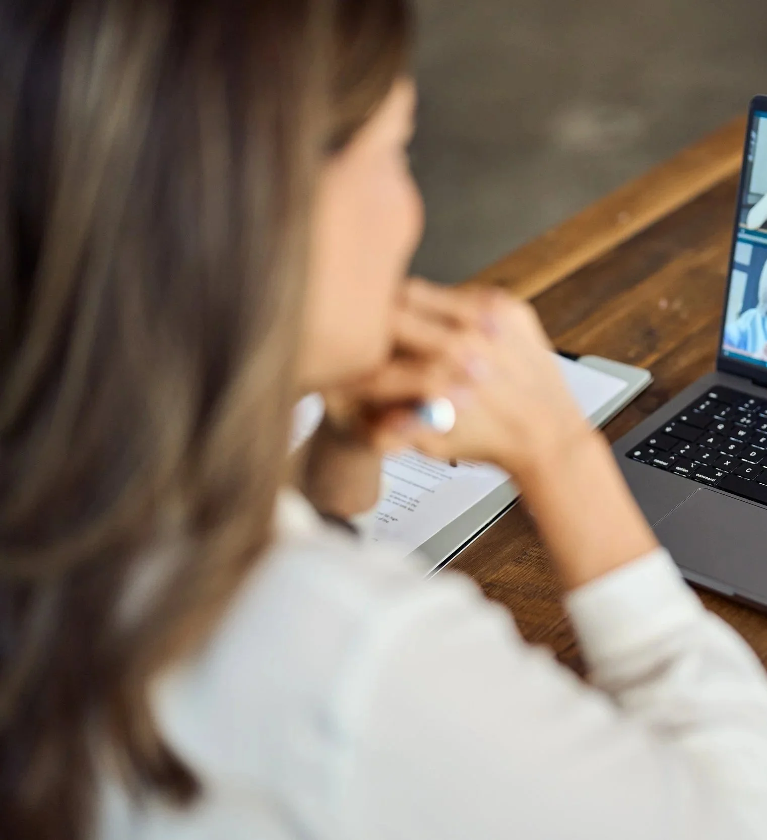 A woman with brown hair is sitting at a wooden table, looking at a laptop screen. She has a notebook or paper in front of her, and appears to be engaged with her work or a video call.