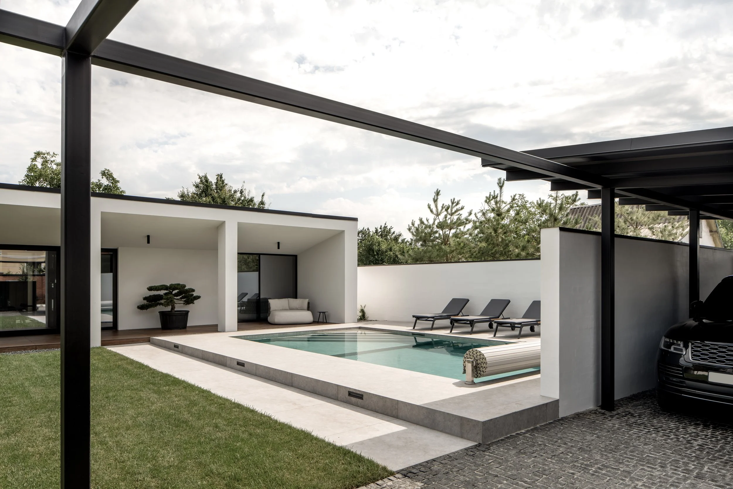 Modern backyard with a swimming pool, lounge chairs, patio furniture, and a minimalist house with large glass doors, surrounded by a white wall and trees.