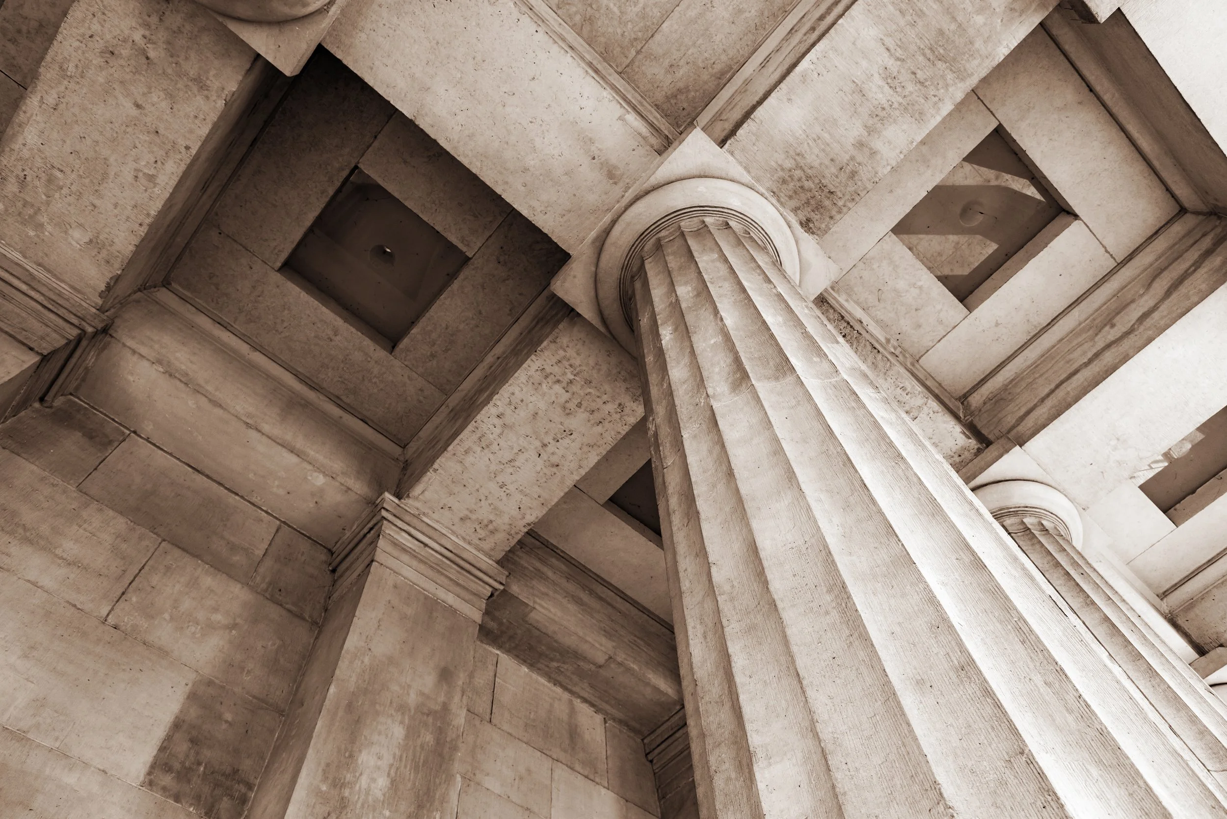 Looking up at a classical architectural ceiling with a large column in the center and intricate molding details, made of light-colored stone.