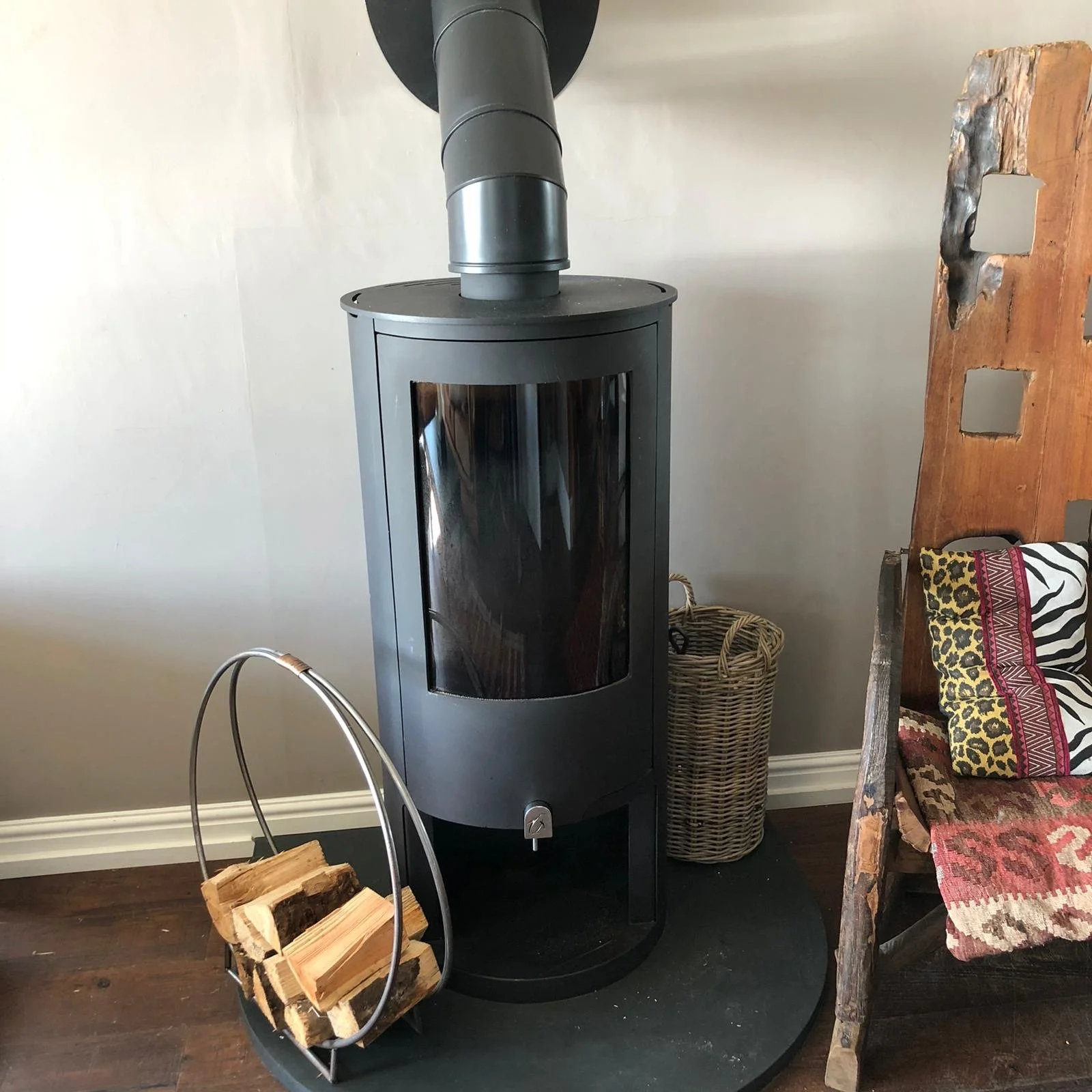A modern wood stove with a glass door, a metal pipe leading upwards, next to a basket for firewood, positioned against a light-colored wall in a cozy living space.