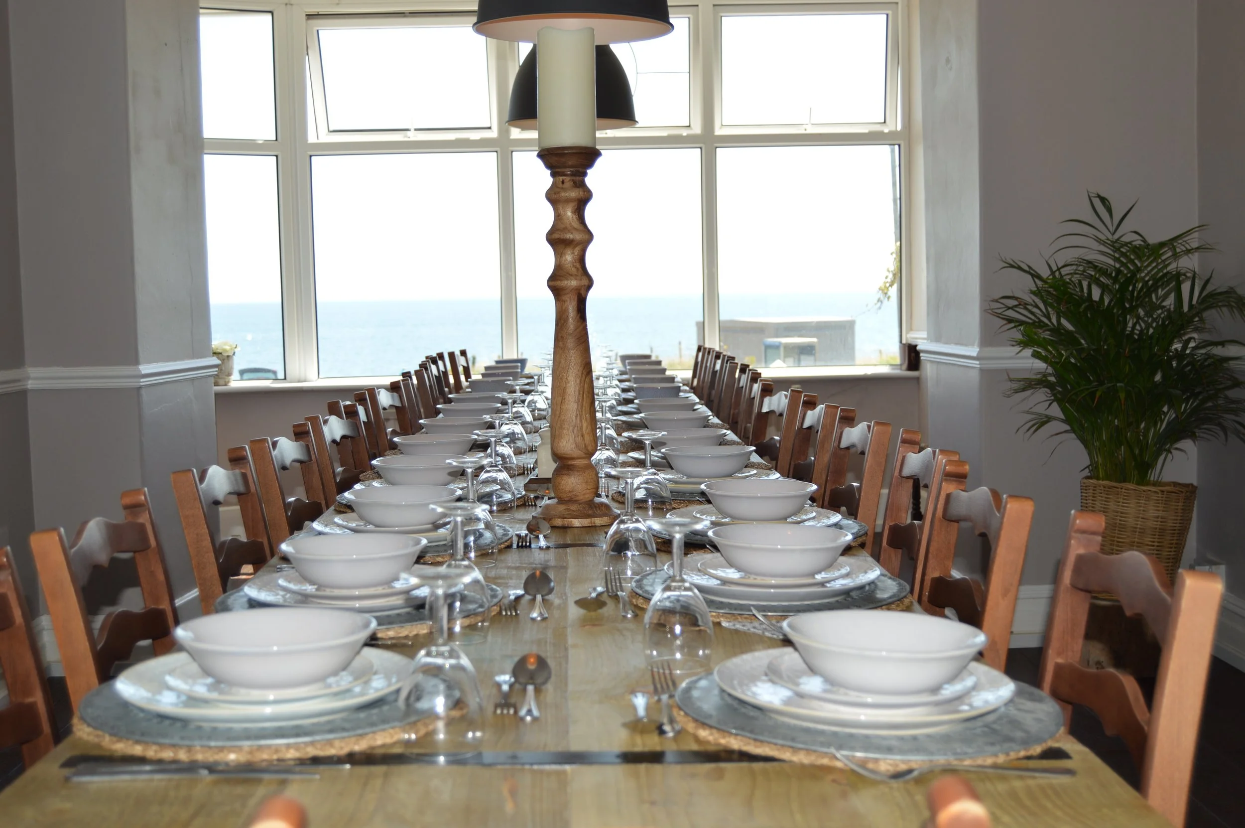 A long dining table set with white bowls, glasses, and silverware, in a room with large windows looking out to the sea, decorated with a potted plant on the side.