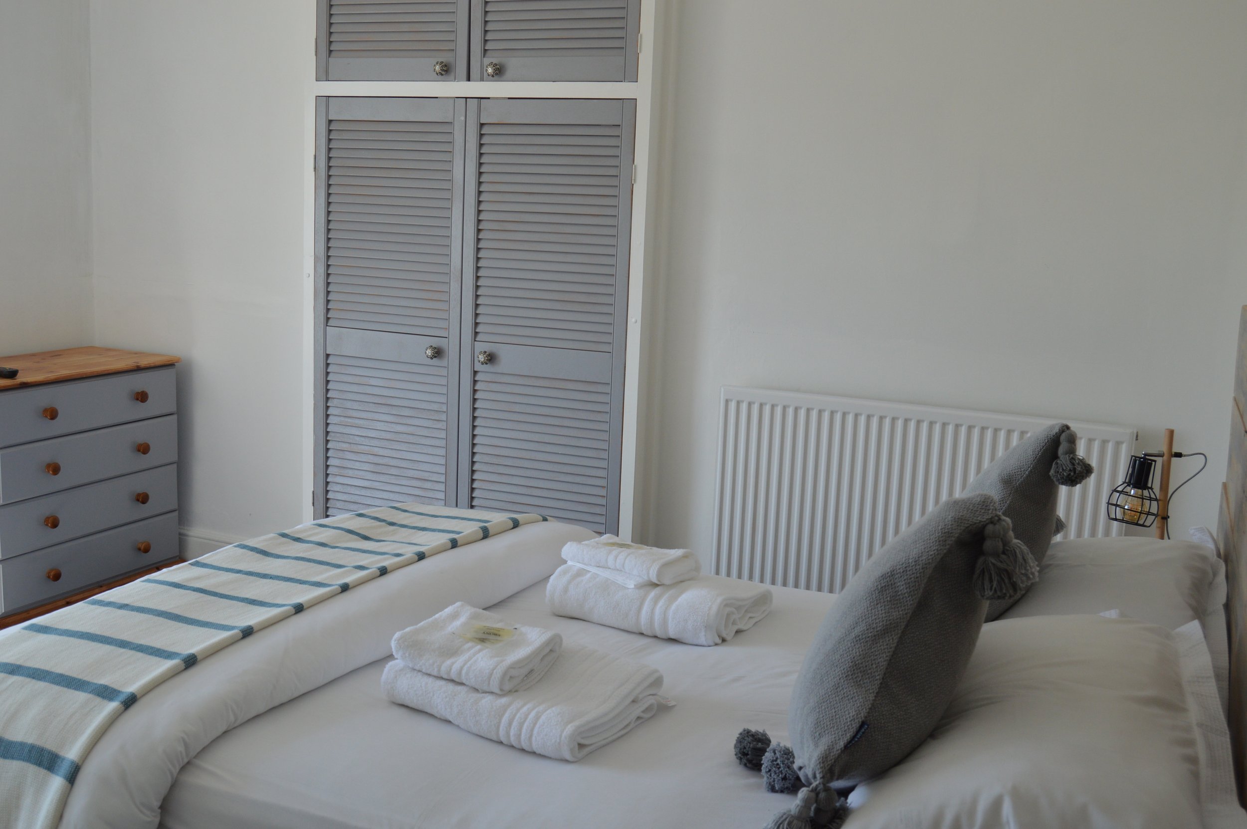A neatly made bed with white linens, a striped blanket, and folded towels, in a bedroom with grey furniture, including a dresser and a closet, and a bedside table with a lighted lamp.