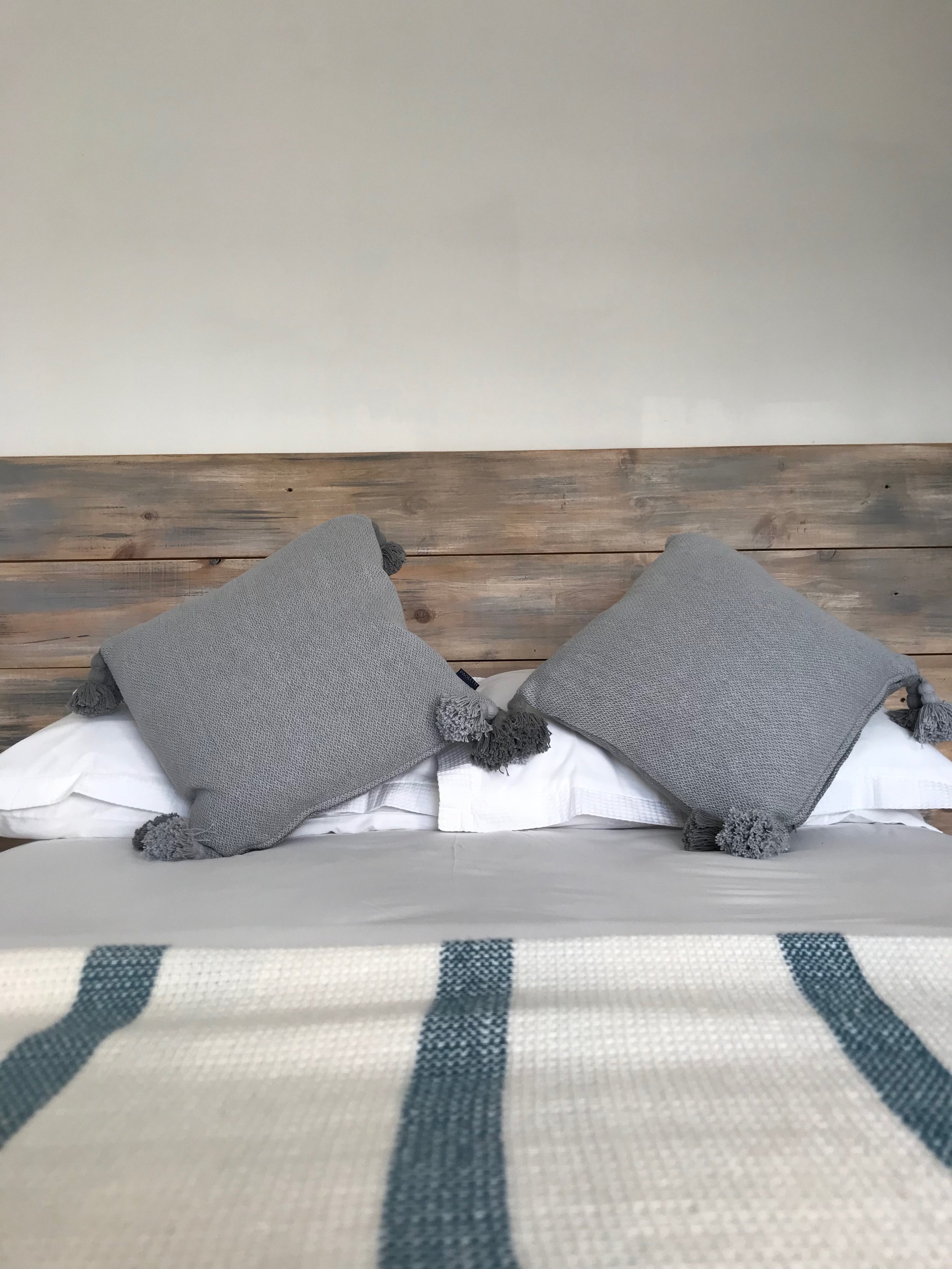 View from above of a bed with three gray pillows and a white and blue striped blanket, with a wooden headboard and a plain beige wall in the background.
