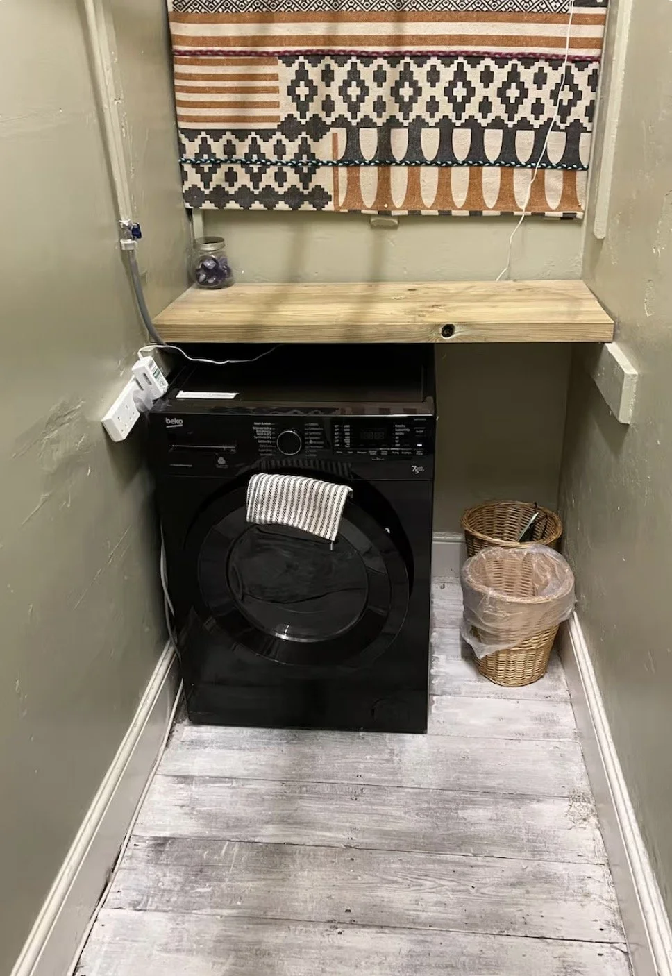 Small laundry room with a black front-loading washing machine below a wooden countertop, a small basket and a trash can next to it, a power outlet with a plug, a jar with purple balls, and patterned curtains on the window.