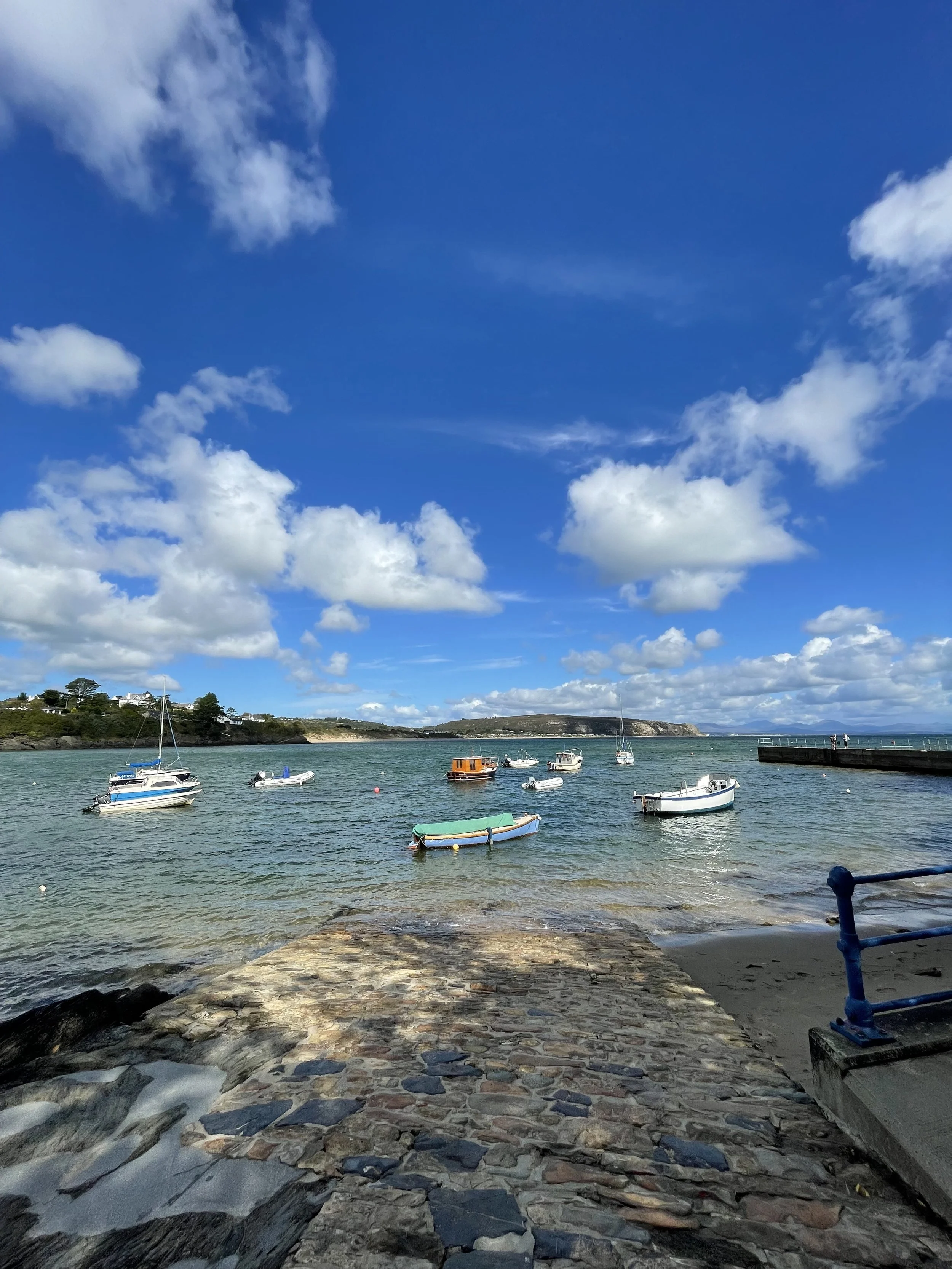 A scenic view of a harbor with small boats anchored in the water, a stone pathway in the foreground, and a blue sky with scattered white clouds overhead. Hills with houses are visible in the background.