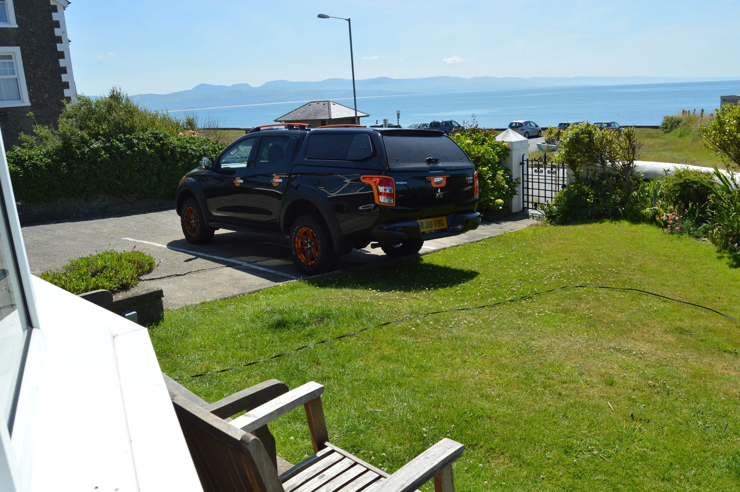 A black Mitsubishi pickup truck with orange accents parked near a garden and a view of the ocean, with cars parked near a small pavilion, trees, and mountains in the background.