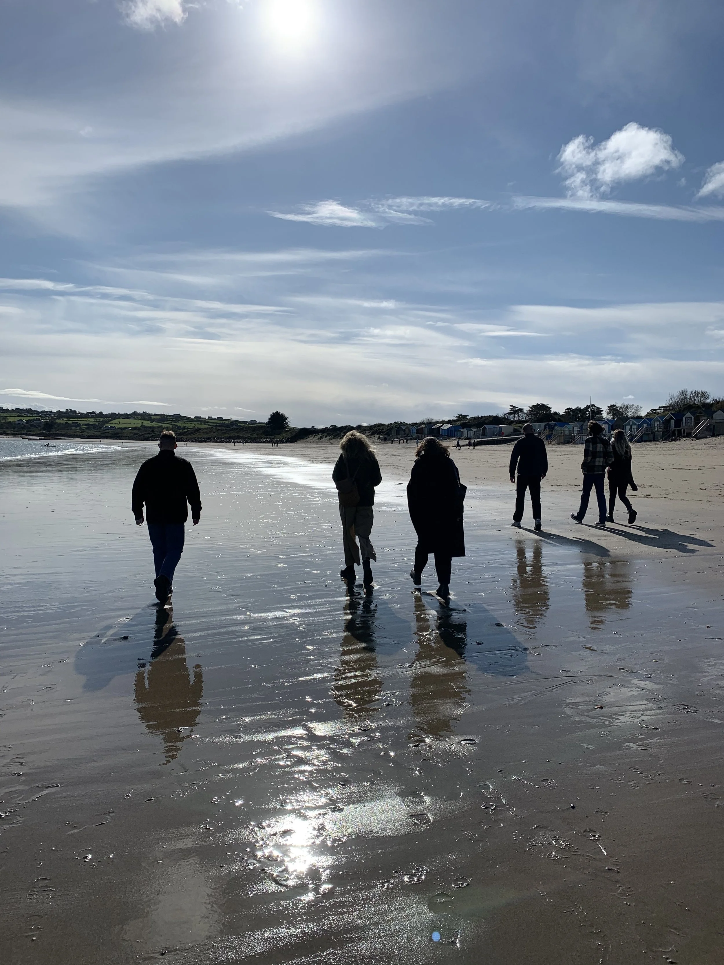 People walking along a beach with the ocean on their left, lifeguard huts on the right, and a partly cloudy sky overhead, with sunlight reflecting on wet sand.