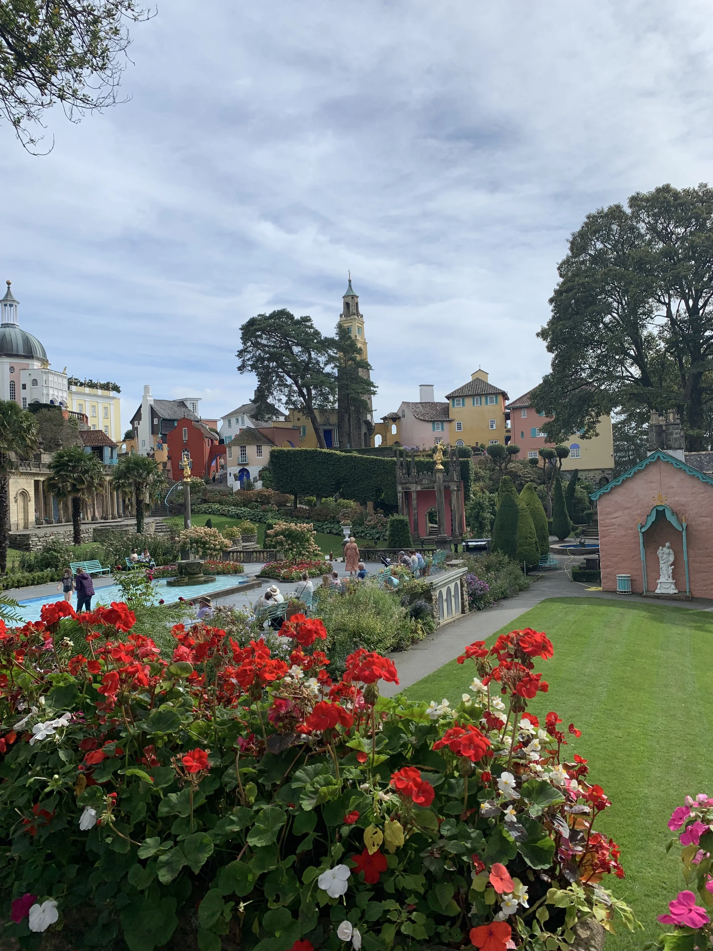 Colorful European-style village with pastel houses, a church clock tower, lush greenery, and vibrant flower beds, under a cloudy sky.