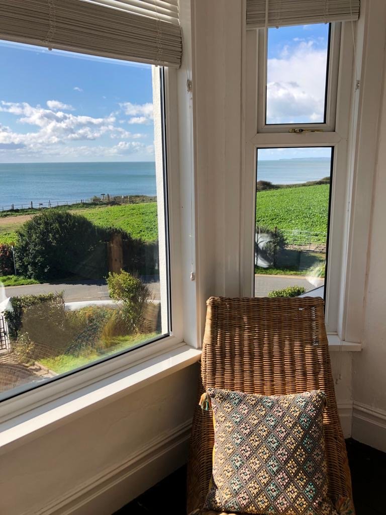 View from a window showing the ocean, green grassy fields, and a partly cloudy sky. Inside the room, there is a wicker chair with a patterned cushion.